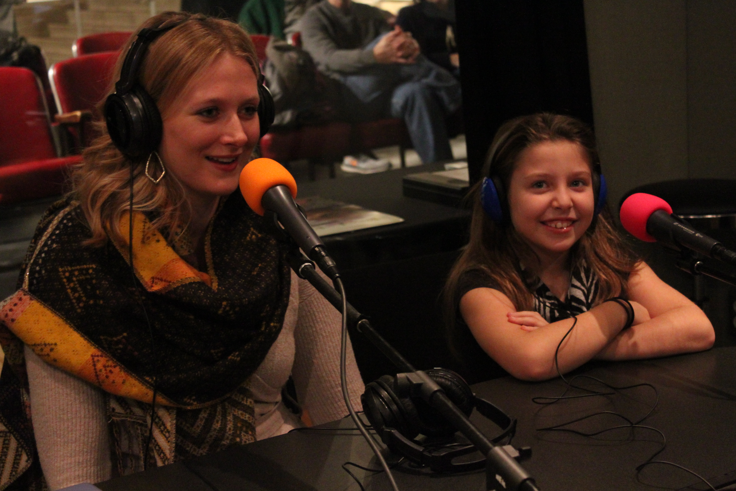 A youth and an adult wearing headphones speaking into microphones in a radio studio