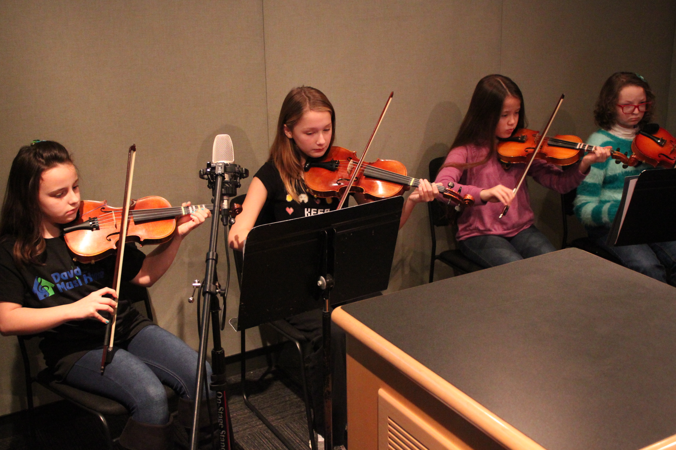 Four youth playing violins looking at sheet music on music stands in a radio studio