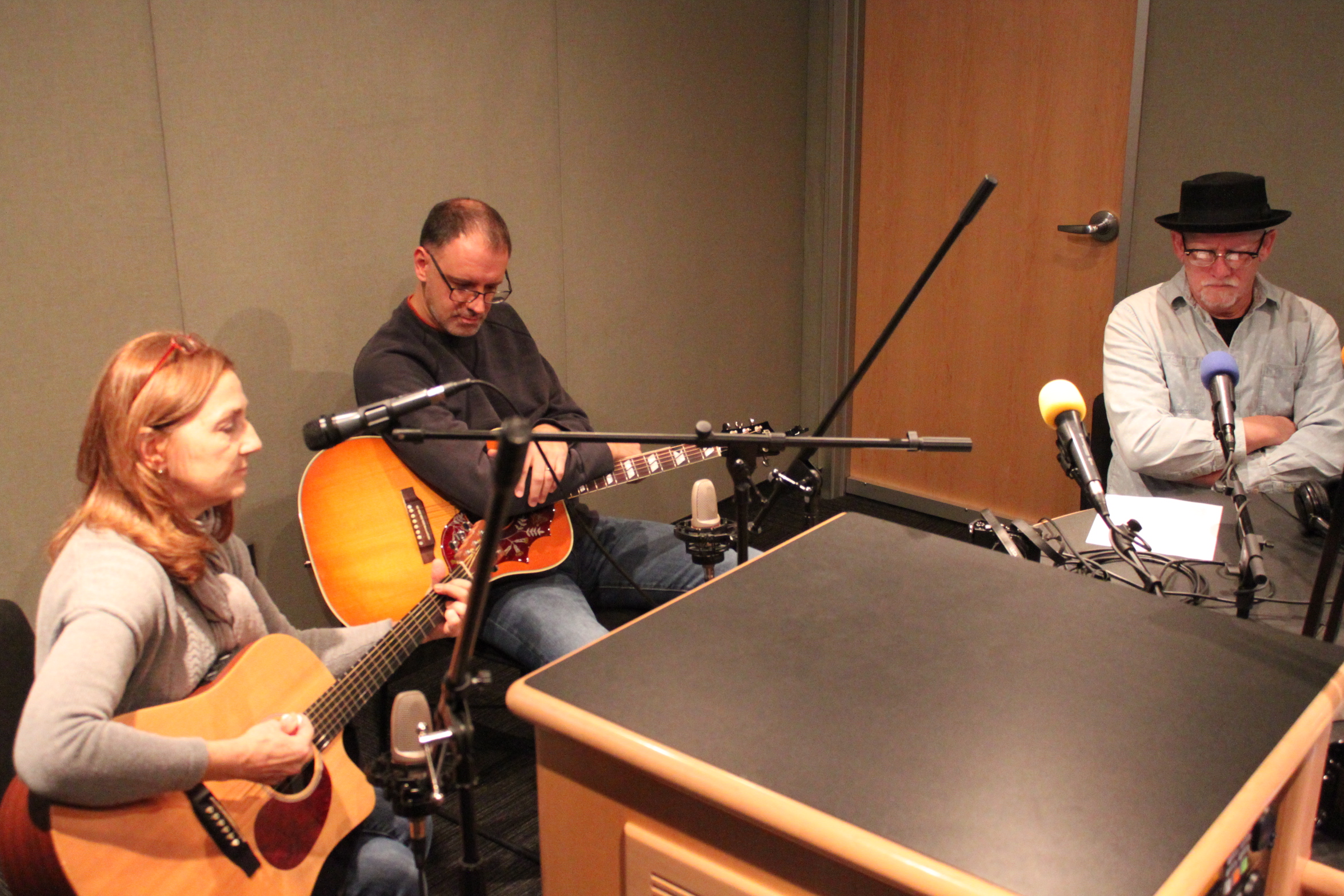 Two adults playing acoustic guitars and an adult sitting behind a microphone in a radio studio