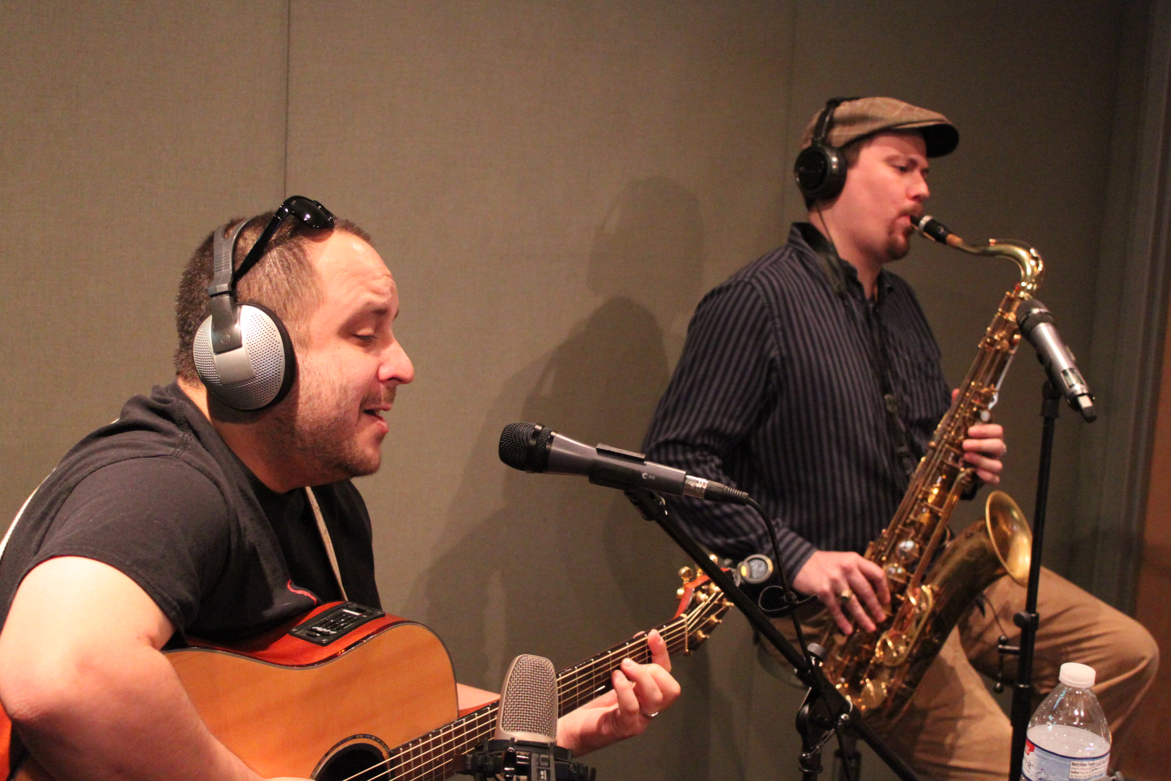 An adult wearing headphones playing an acoustic guitar and an adult wearing headphones playing a tenor saxophone in a radio studio