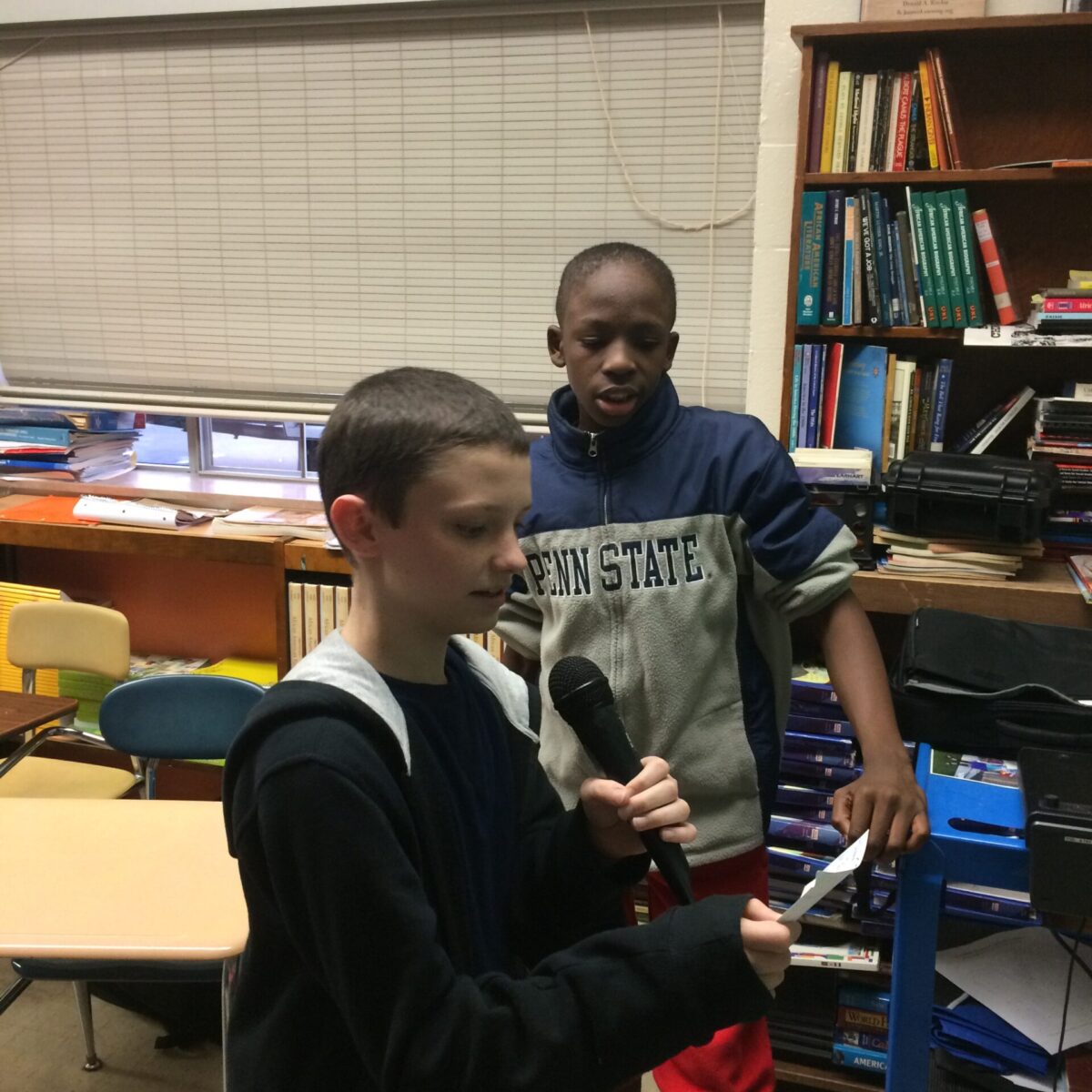 Two youth standing in a classroom, one holding a microphone and looking down at a piece of paper