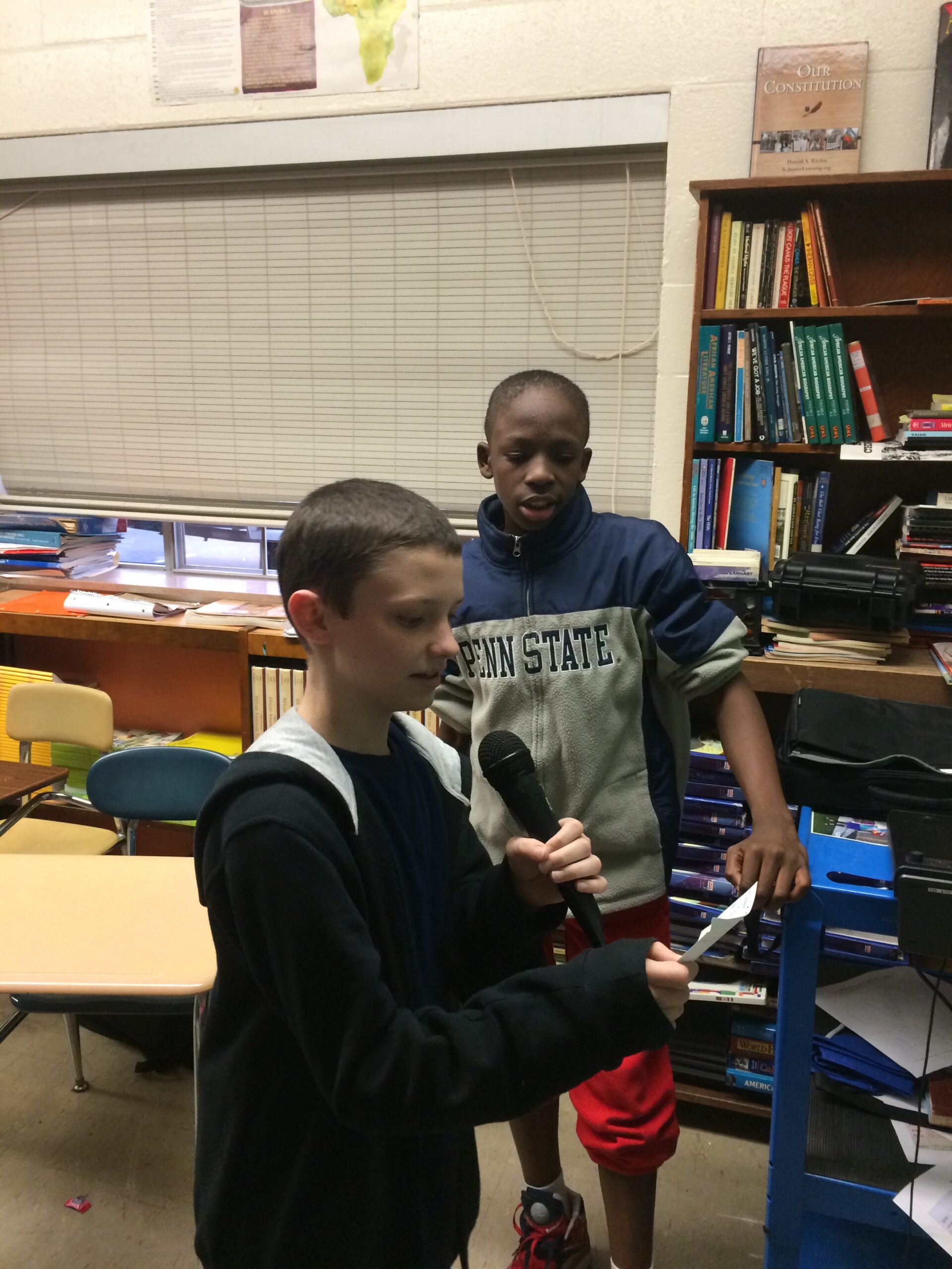 Two youth standing in a classroom, one holding a microphone and looking down at a piece of paper