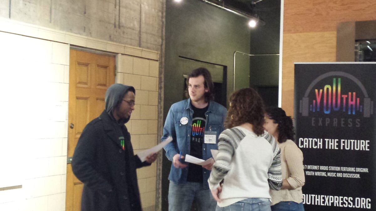 Three teens holding pieces of paper and an adult standing in a circle in front of a radio studio