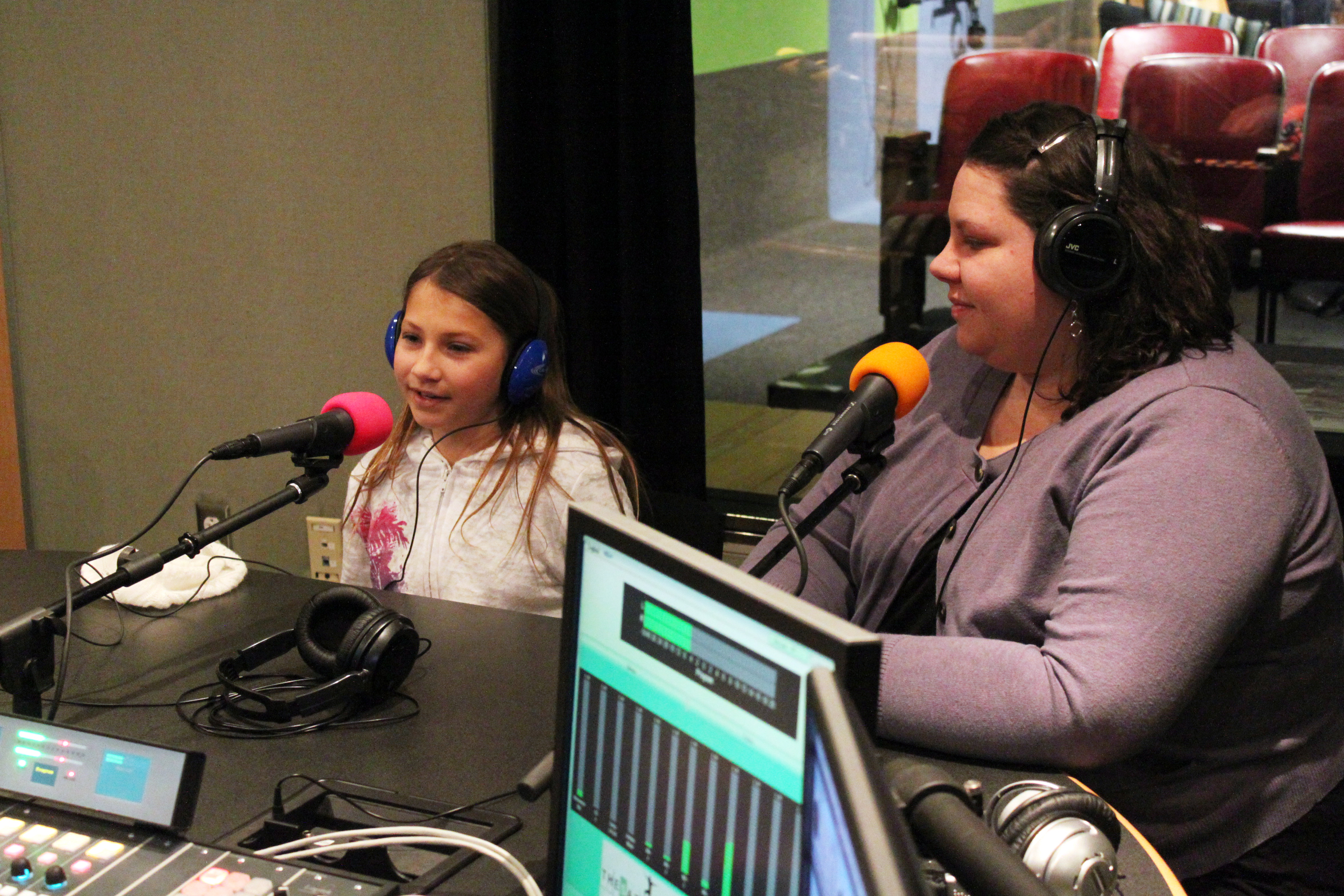 An adult and a youth wearing headphones speaking into microphones in a radio studio