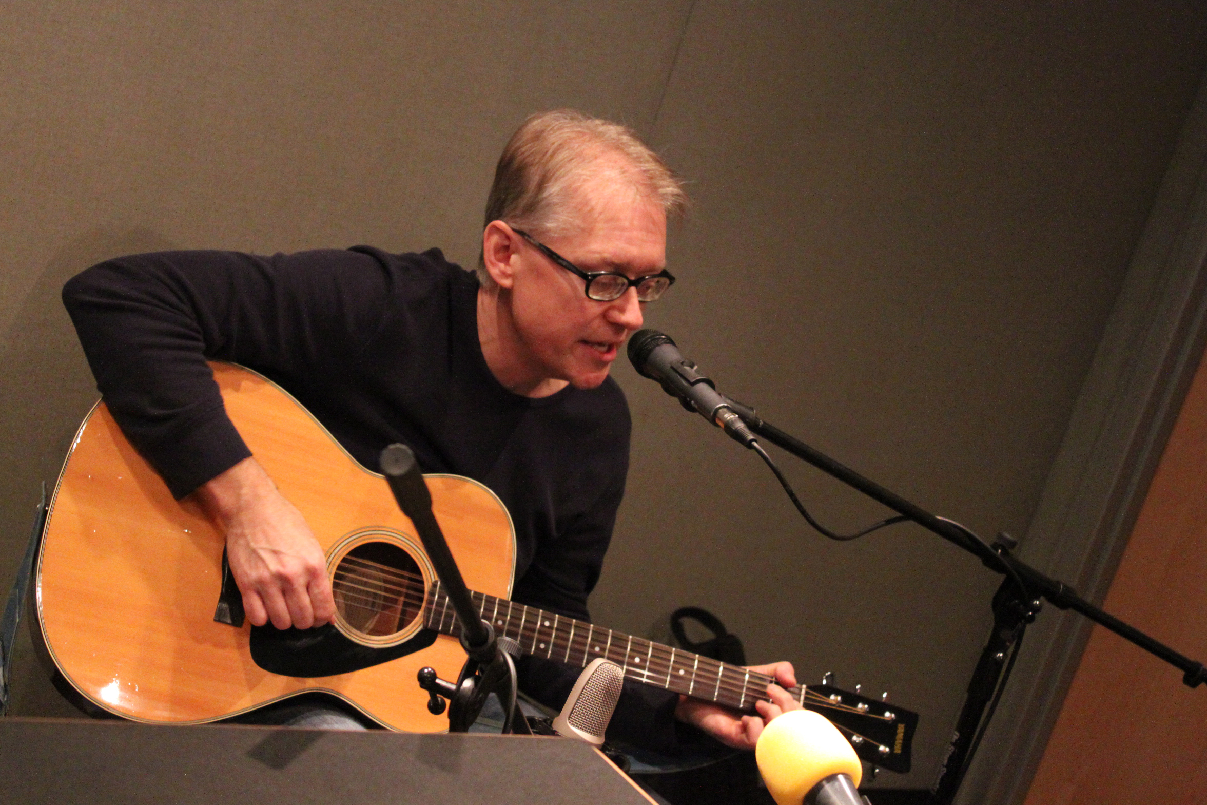 An adult playing acoustic guitar and singing into a microphone in a radio studio