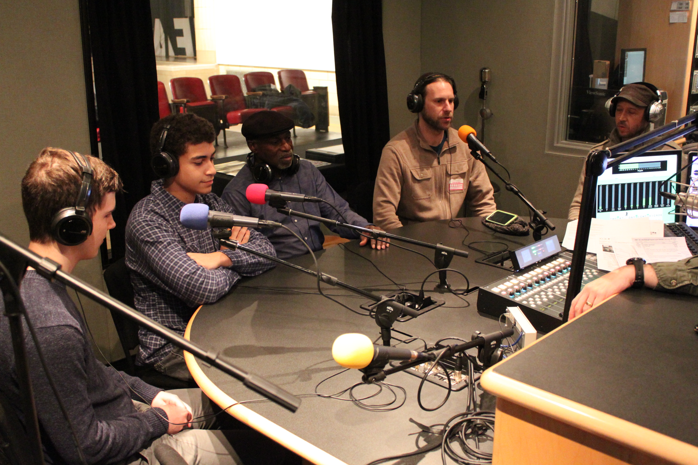 Two teens and three adults wearing headphones speaking into microphones being interviewed in a radio studio