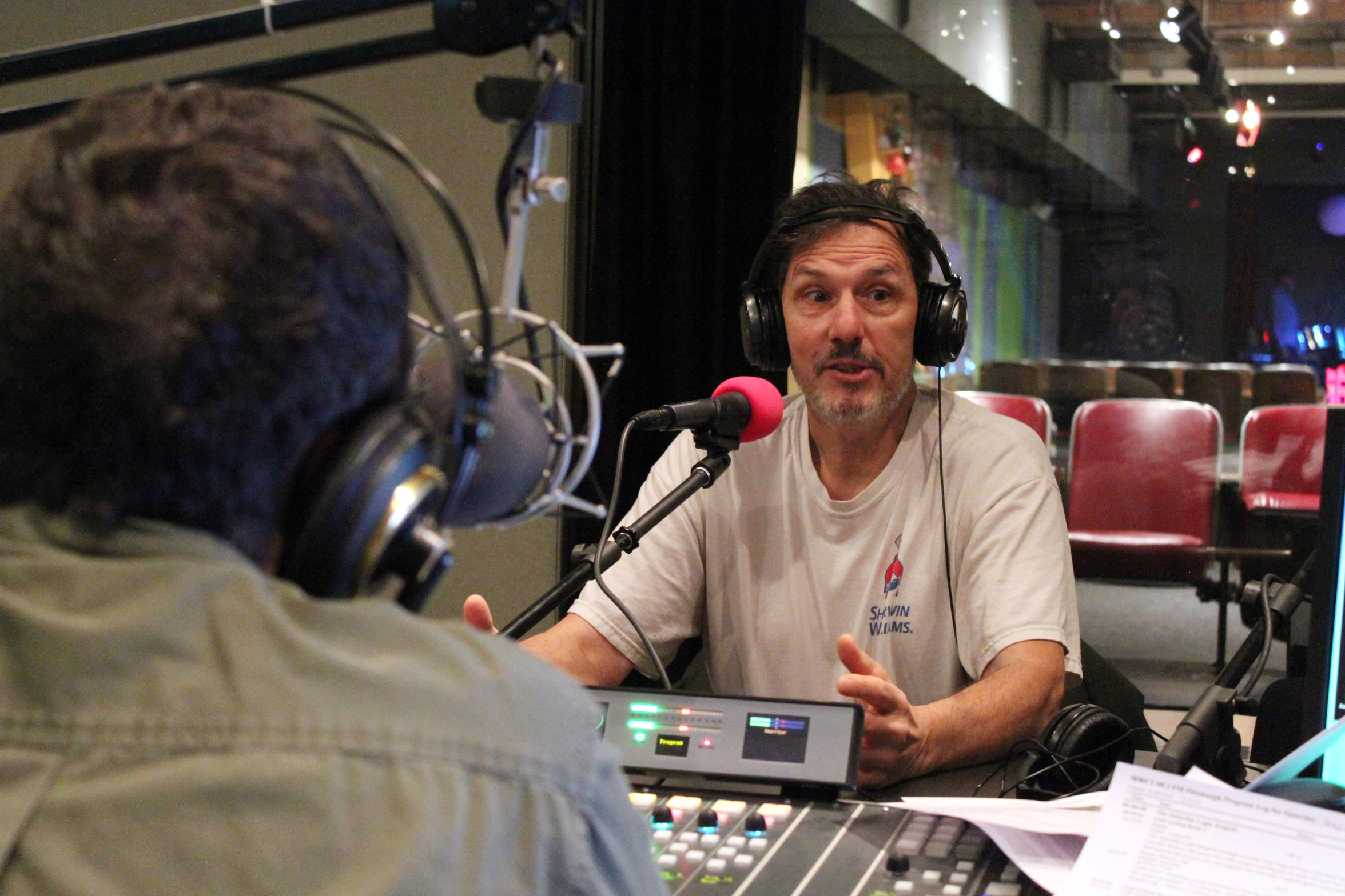 An adult wearing headphones speaking into a microphone being interviewed by an adult wearing headphones sitting behind a microphone and a mixing board in a radio studio