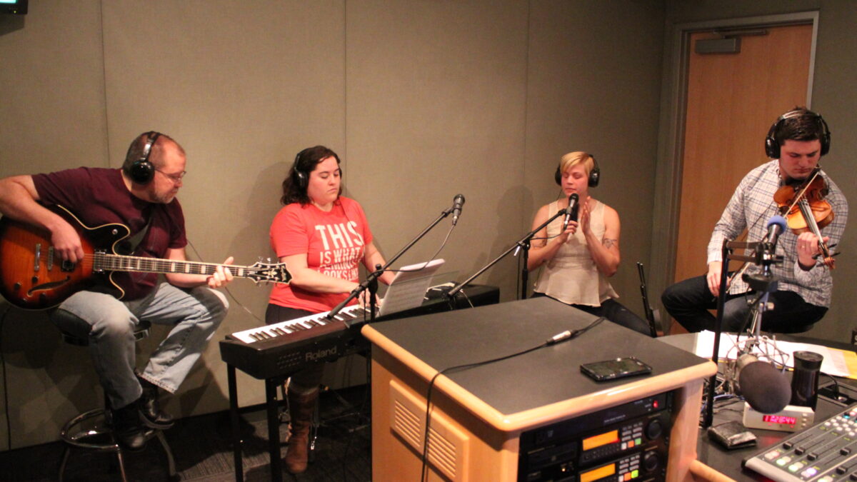 IMG_0633 An adult wearing headphones playing acoustic guitar, an adult wearing headphones playing a piano keyboard, an adult wearing headphones playing a shaker, and an adult wearing headphones playing a violin in a radio studio