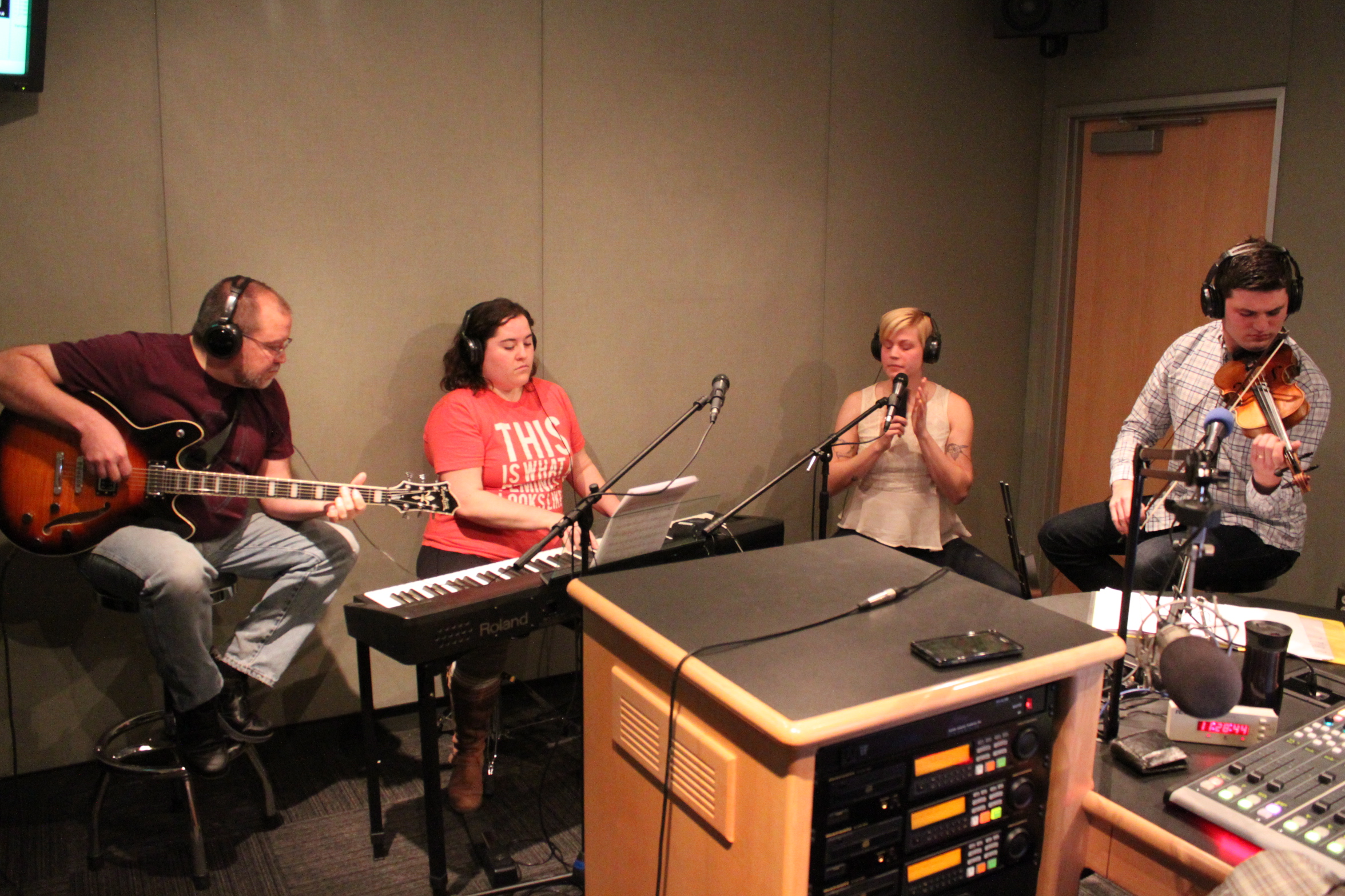 An adult wearing headphones playing acoustic guitar, an adult wearing headphones playing a piano keyboard, an adult wearing headphones playing a shaker, and an adult wearing headphones playing a violin in a radio studio