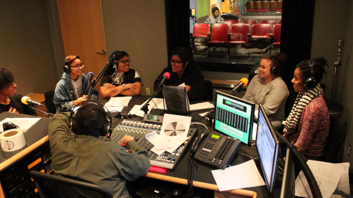 Six teens wearing headphones speaking into microphones and an adult wearing headphones sitting behind a mixing board in a radio studio
