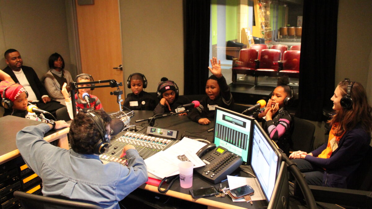 Six youth and an adult wearing headphones speaking into microphones being interviewed by an adult wearing headphones sitting behind a microphone and a mixing board as two adults sit in the background listening in a radio studio