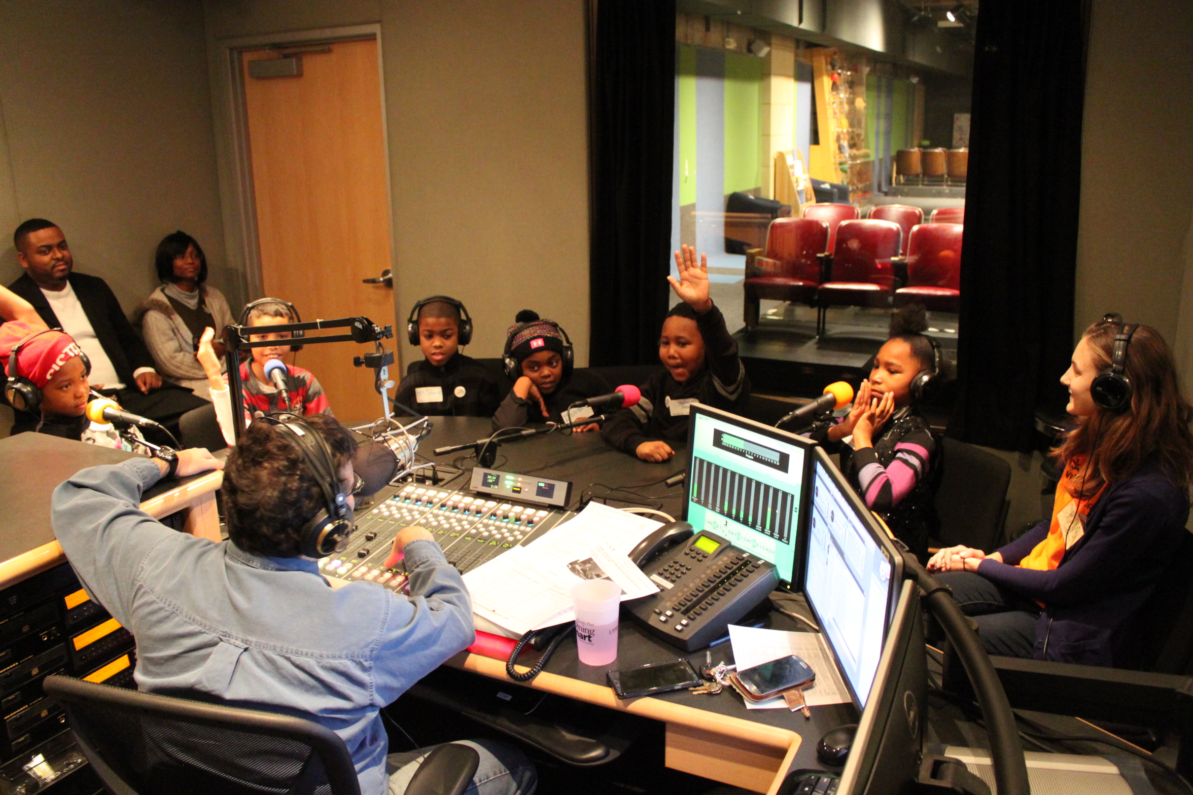 Six youth and an adult wearing headphones speaking into microphones being interviewed by an adult wearing headphones sitting behind a microphone and a mixing board as two adults sit in the background listening in a radio studio