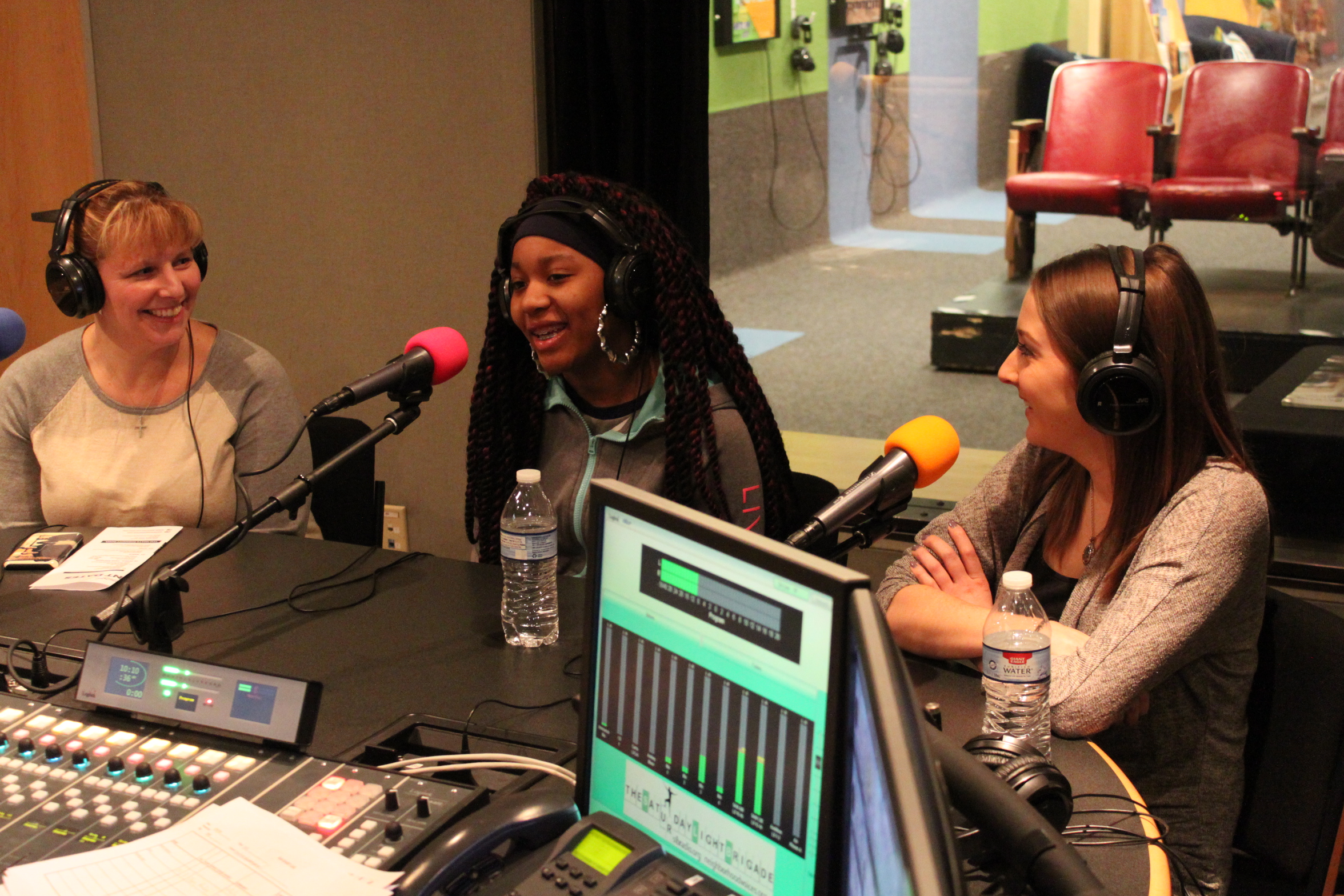 Two adults and a youth wearing headphones speaking into microphones in a radio studio