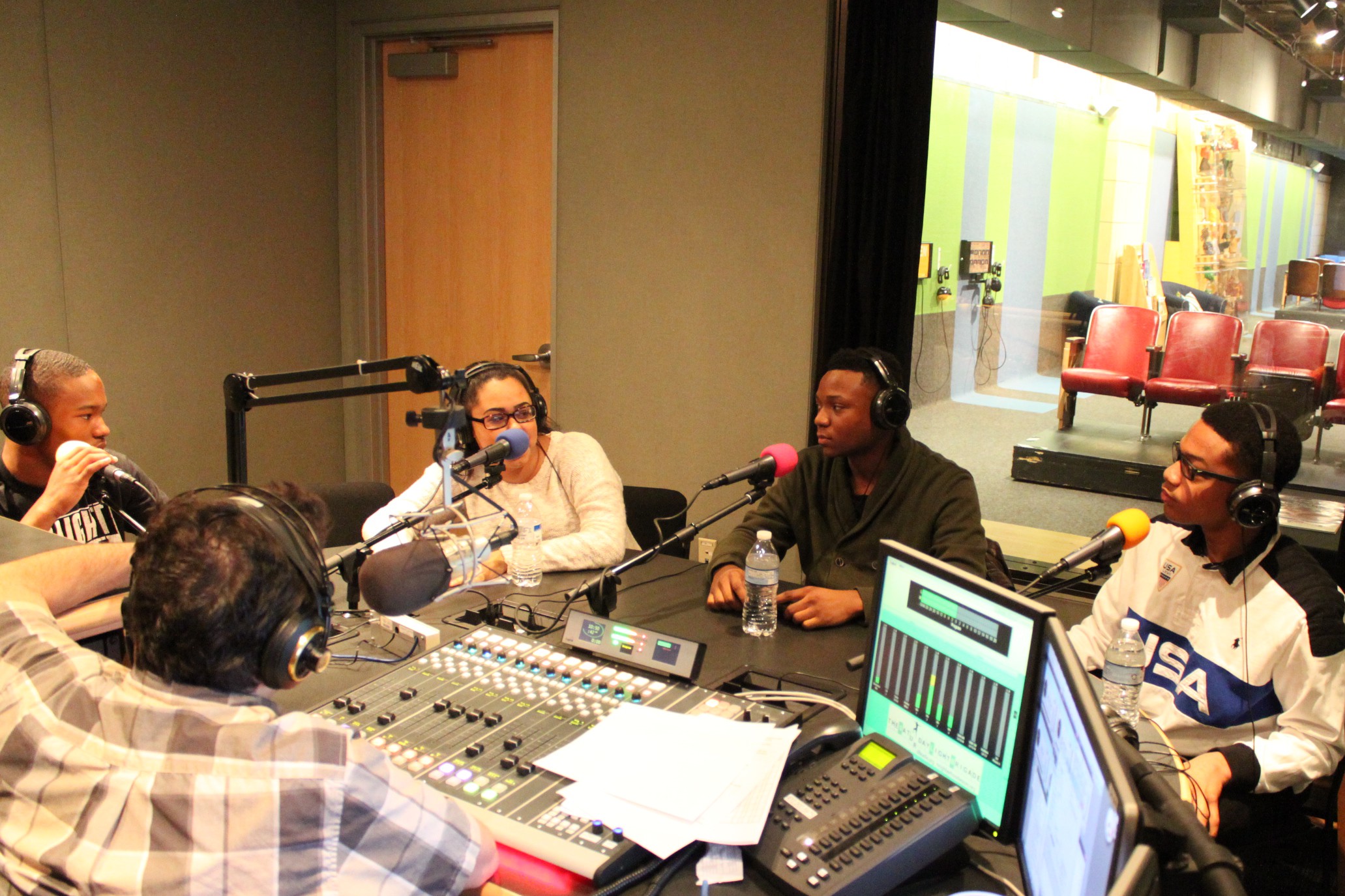 Four teens wearing headphones speaking into microphones being interviewed by an adult wearing headphones sitting behind a microphone and a mixing board in a radio studio