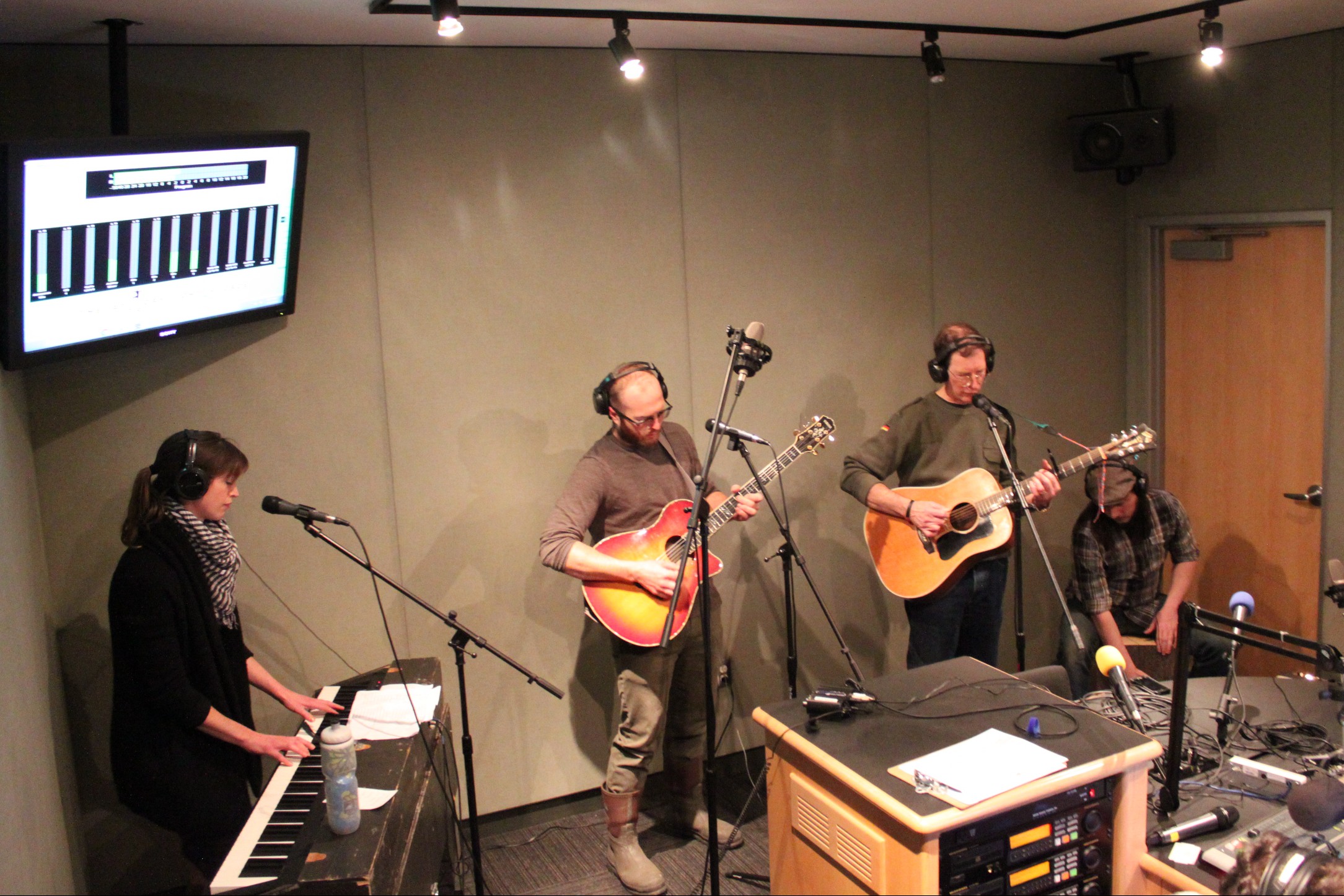 An adult wearing headphones playing a piano keyboard, two adults wearing headphones playing acoustic guitars, and an adult wearing headphones playing a cajon in a radio studio