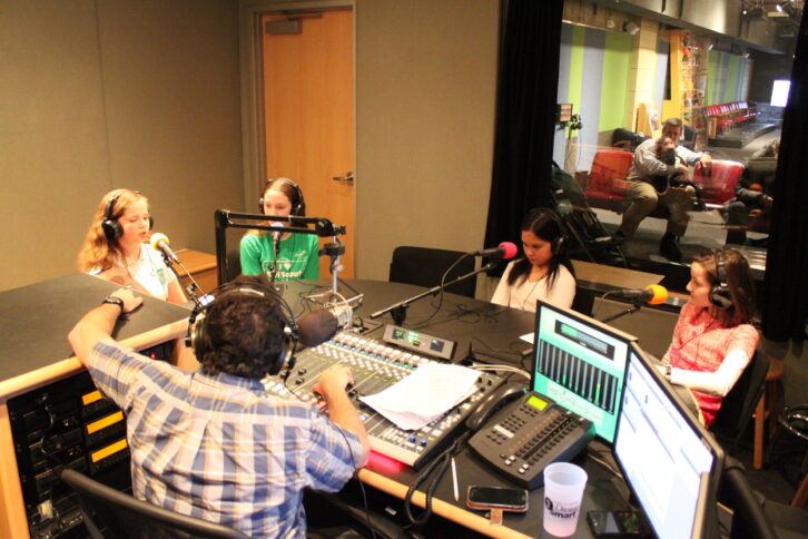 Four youth wearing headphones speaking into microphones being interviewed by an adult wearing headphones sitting behind a microphone and a mixing board in a radio studio