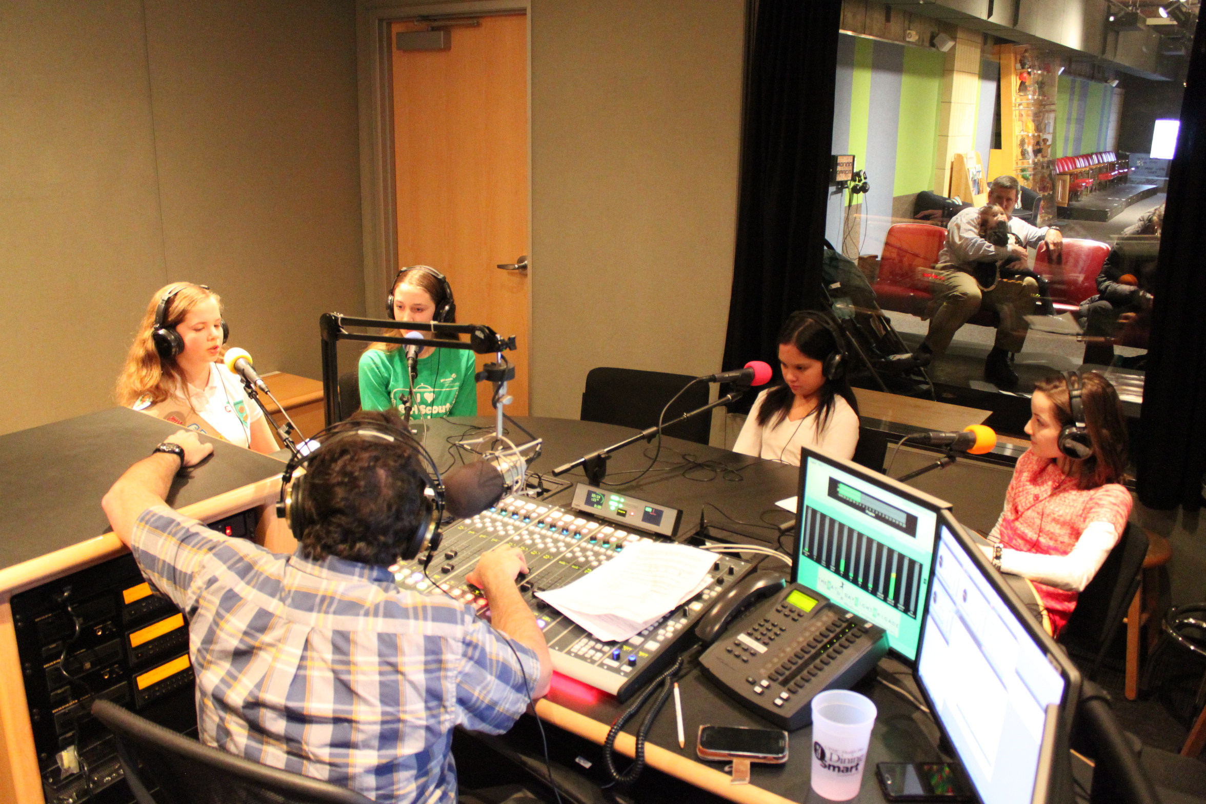 Four youth wearing headphones speaking into microphones being interviewed by an adult wearing headphones sitting behind a microphone and a mixing board in a radio studio