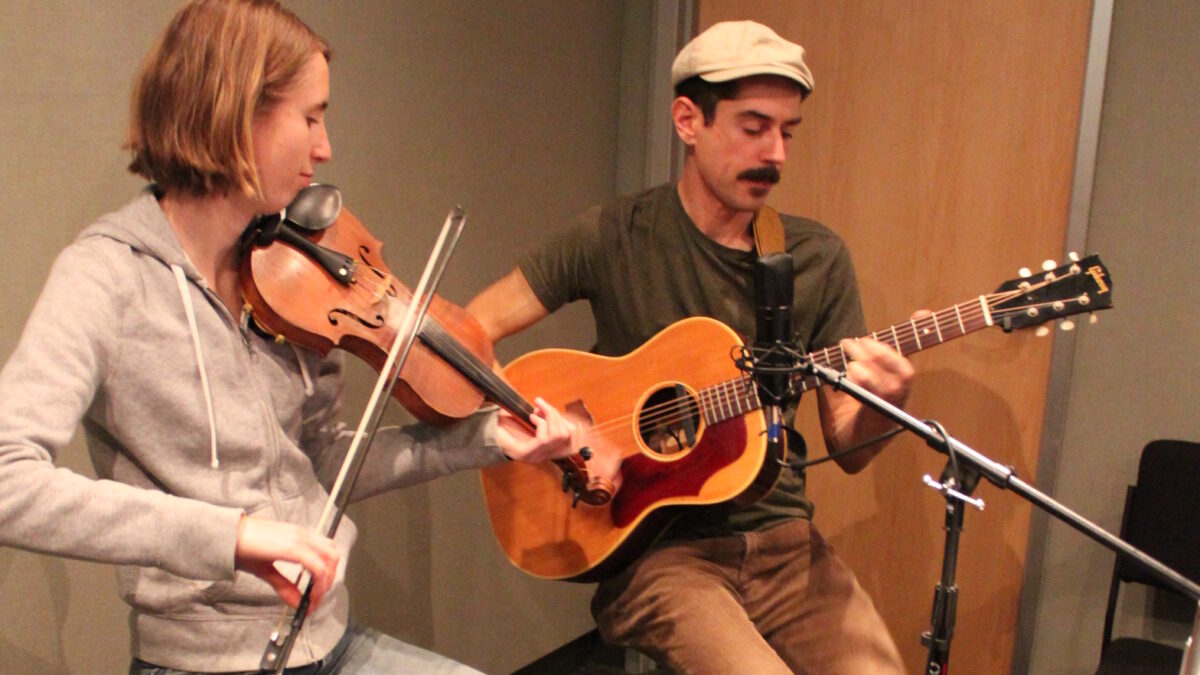 An adult playing fiddle and an adult playing acoustic guitar and singing into a microphone in a radio studio