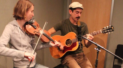 An adult playing fiddle and an adult playing acoustic guitar and singing into a microphone in a radio studio