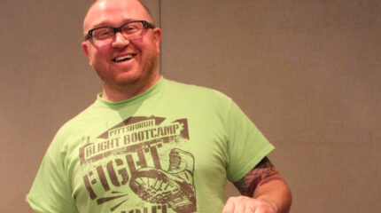 An adult smiling and holding up a framed art piece of a car in front of a home in a radio studio