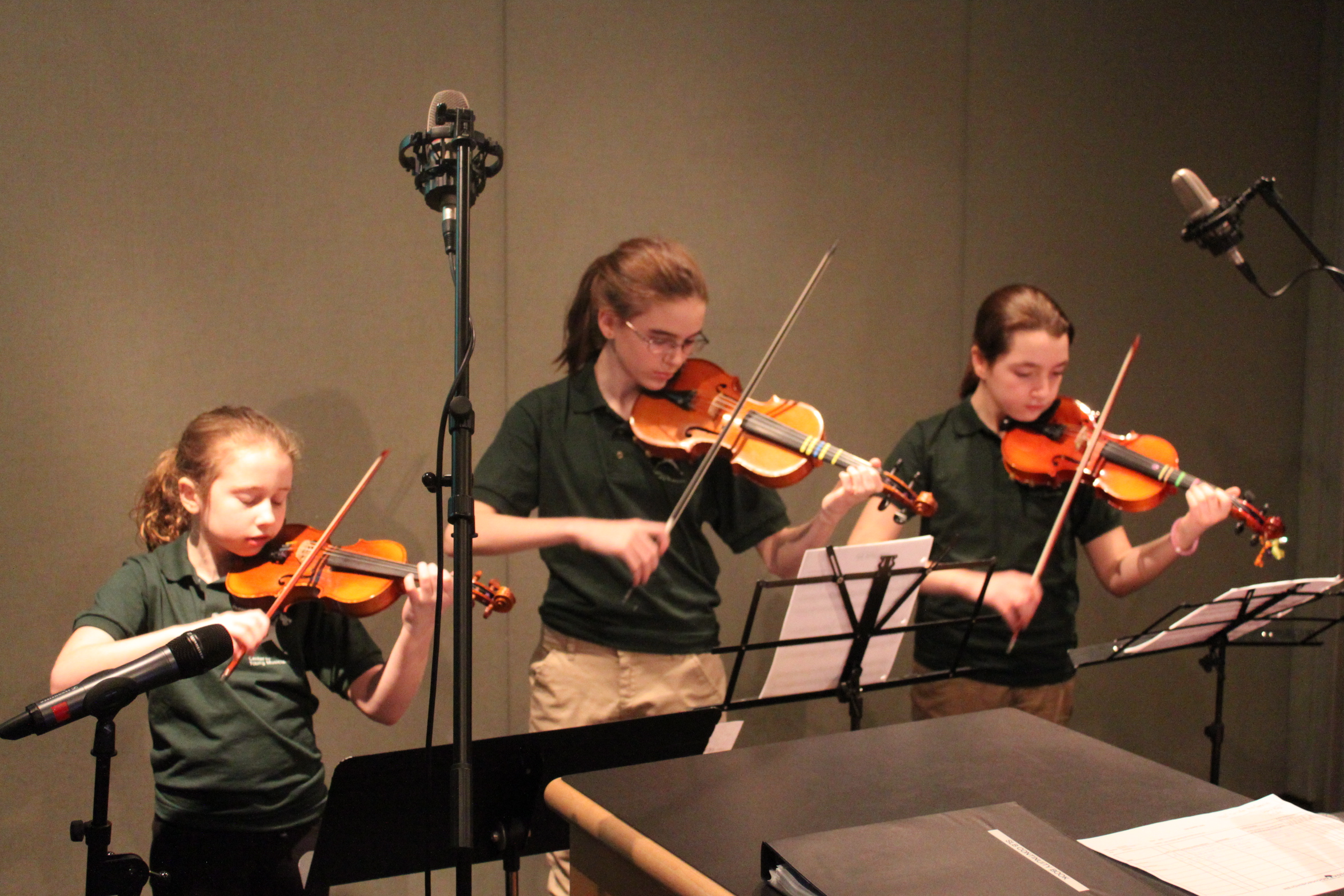 Three youth playing violins looking at sheet music on music stands in a radio studio