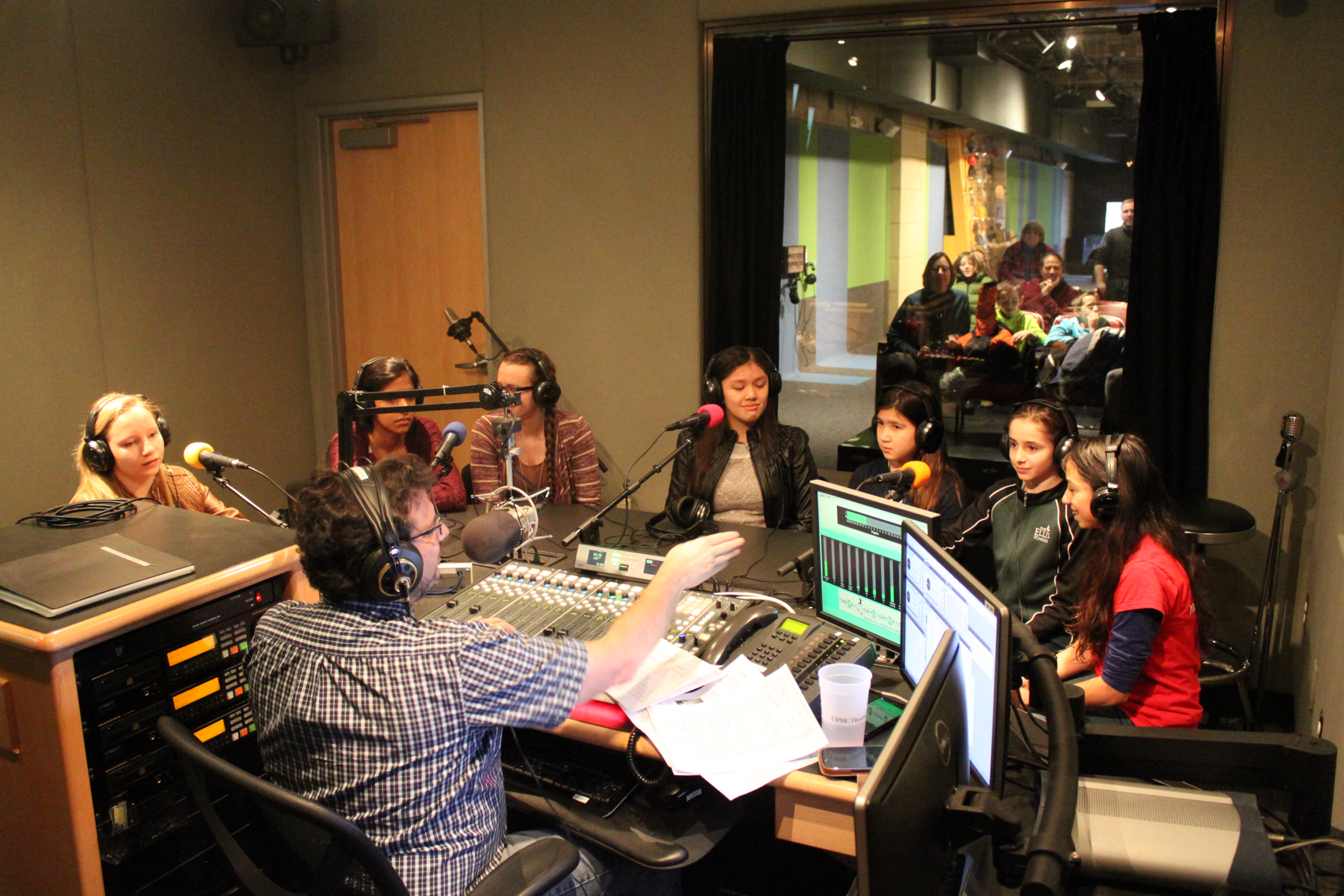 Seven teens wearing headphones speaking into microphones being interviewed by an adult wearing headphones sitting behind a microphone and a mixing board in a radio studio