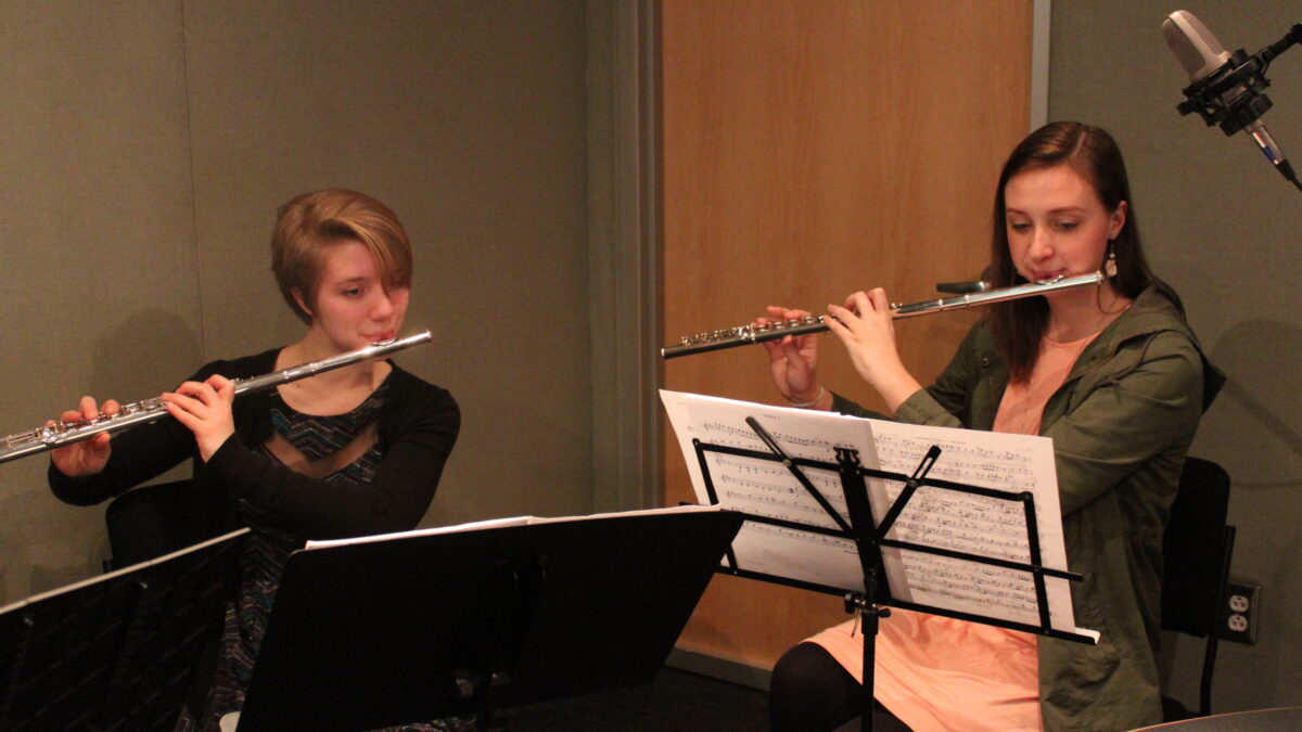 Two teens playing flutes looking at sheet music on music stands in a radio studio