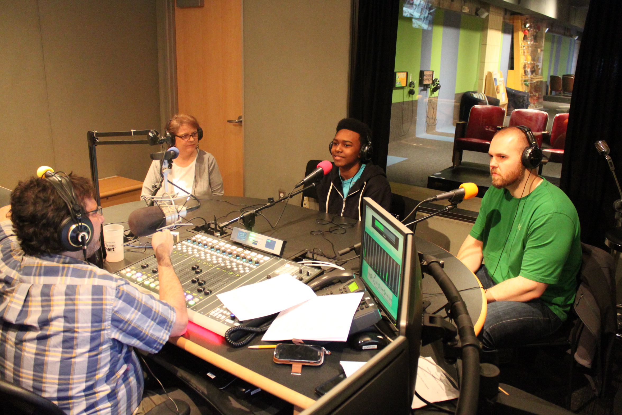 Two adults and a teen wearing headphones speaking into microphones being interviewed by an adult wearing headphones sitting behind a microphone and a mixing board in a radio studio