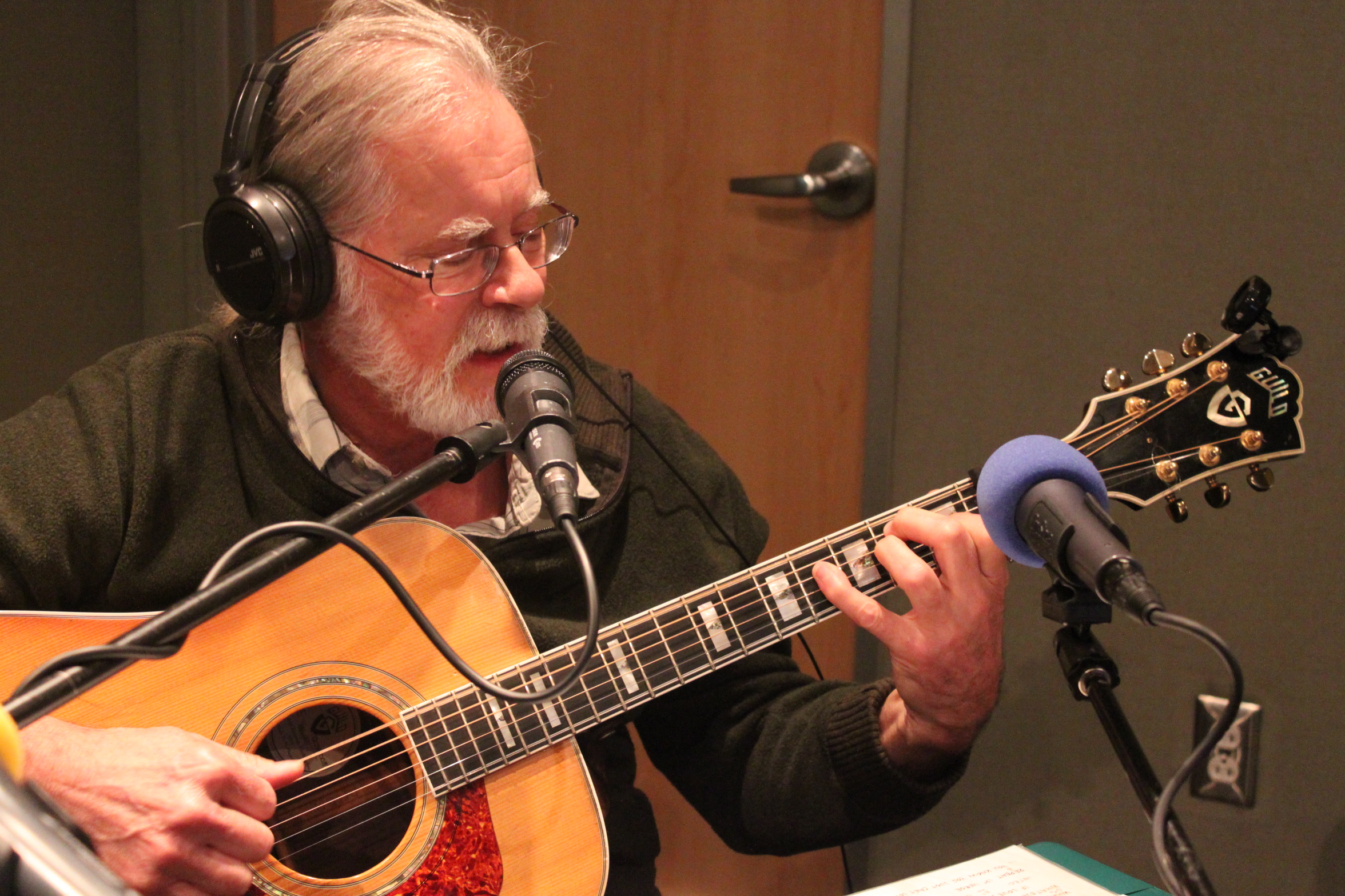 An adult wearing headphones playing acoustic guitar and singing into a microphone in a radio studio