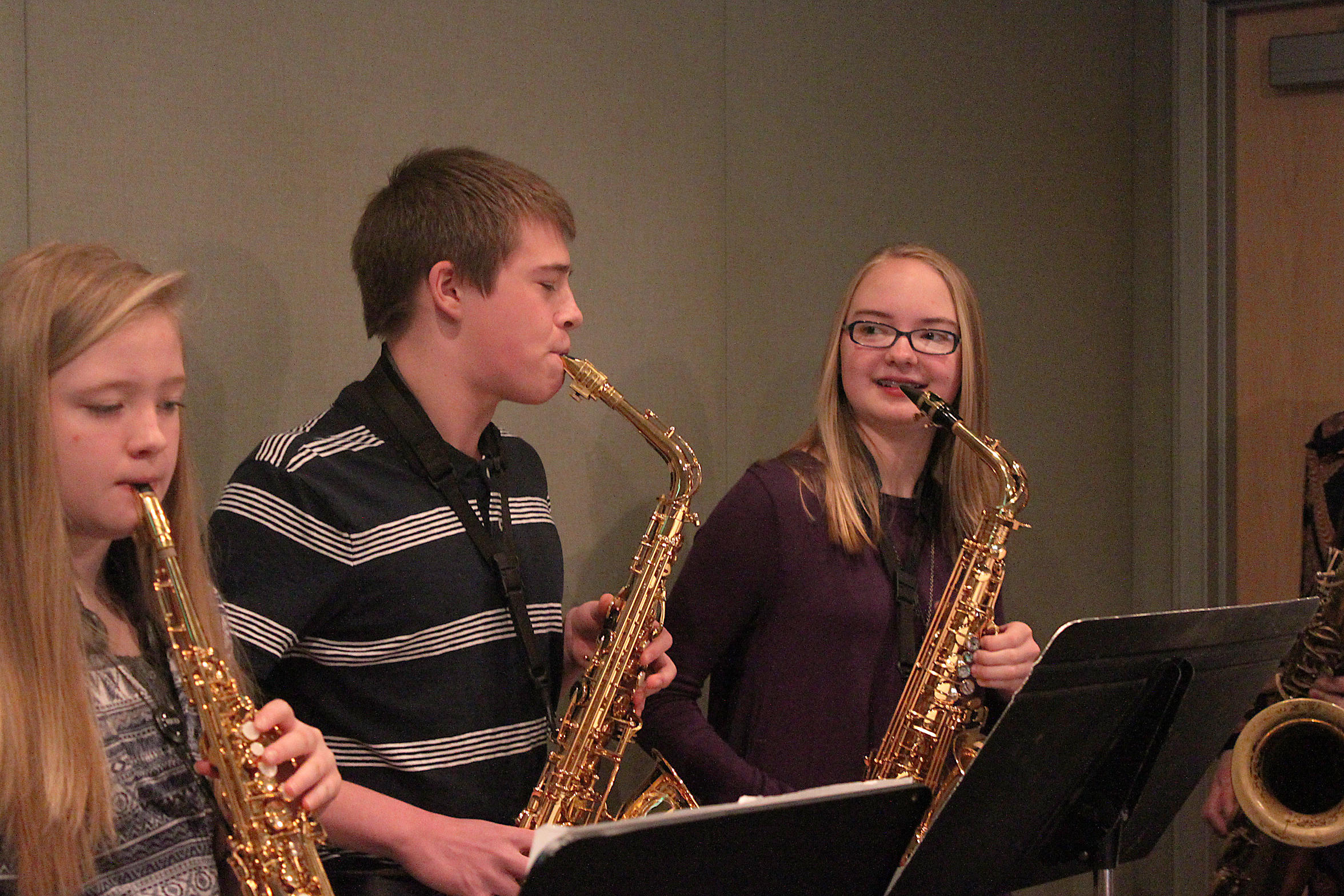 A youth playing soprano saxophone and two youth playing alto saxophones, all looking at sheet music on music stands in a radio studio
