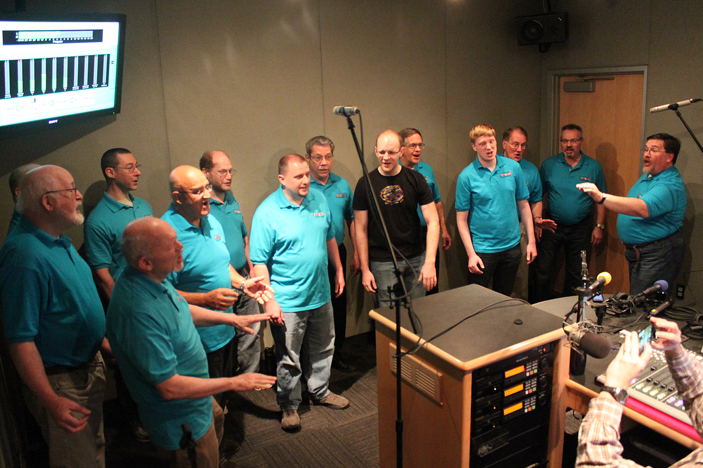 Thirteen adults singing in a choir in a radio studio