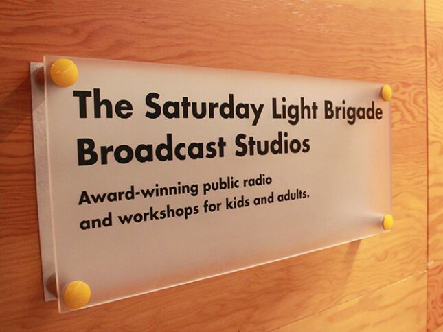 A glass sign on a wooden wall reading "The Saturday Light Brigade Broadcast Studios" and "Award-Winning public radio and workshops for kids and adults."