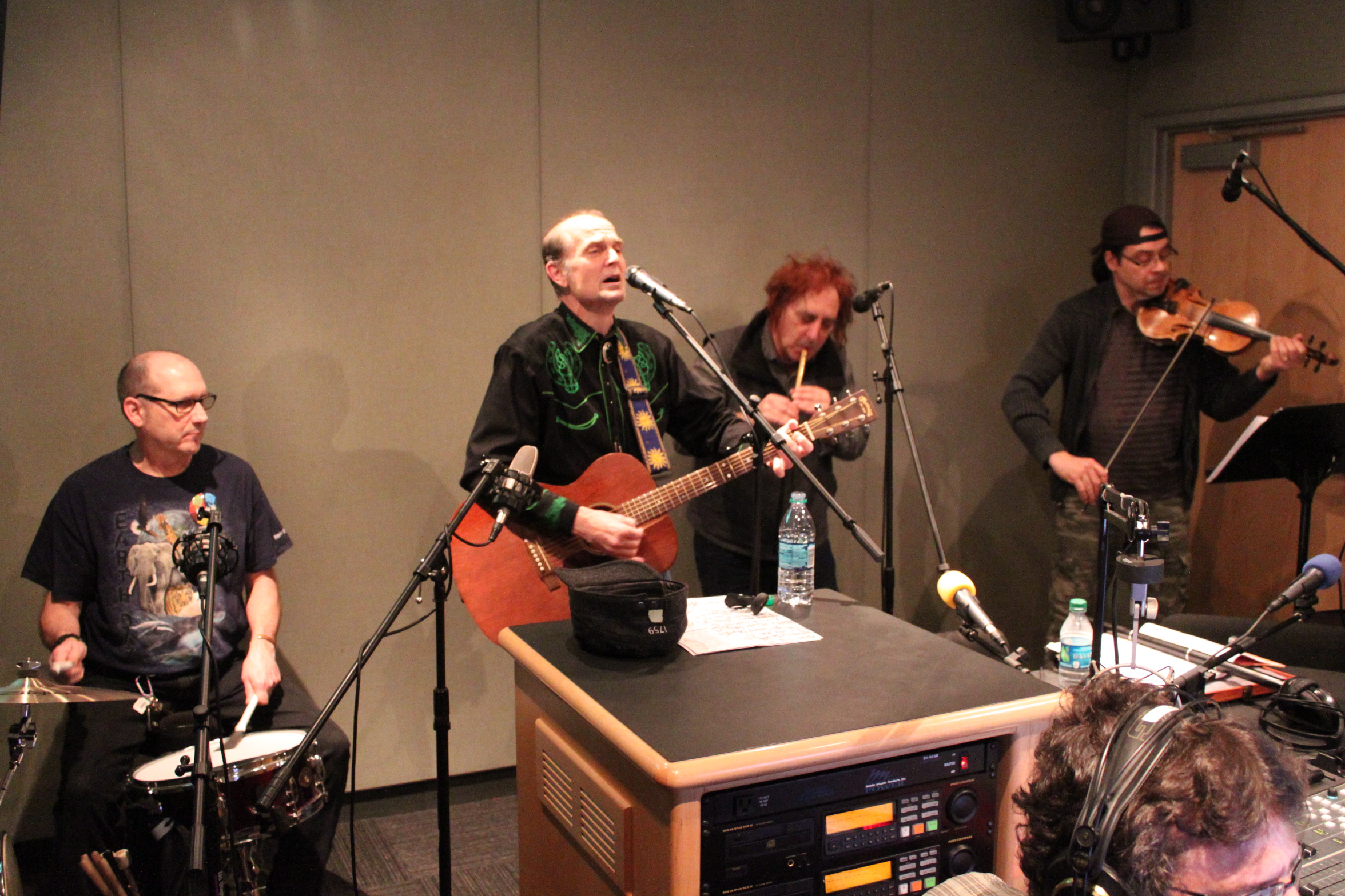An adult playing a drum set, an adult playing acoustic guitar and singing into a microphone, an adult playing whistle, and an adult paying a fiddle in a radio studio