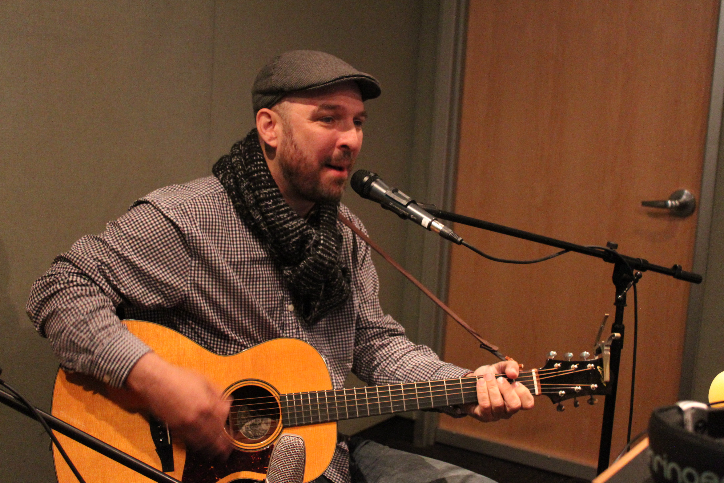 An adult playing acoustic guitar and singing into a microphone in a radio studio