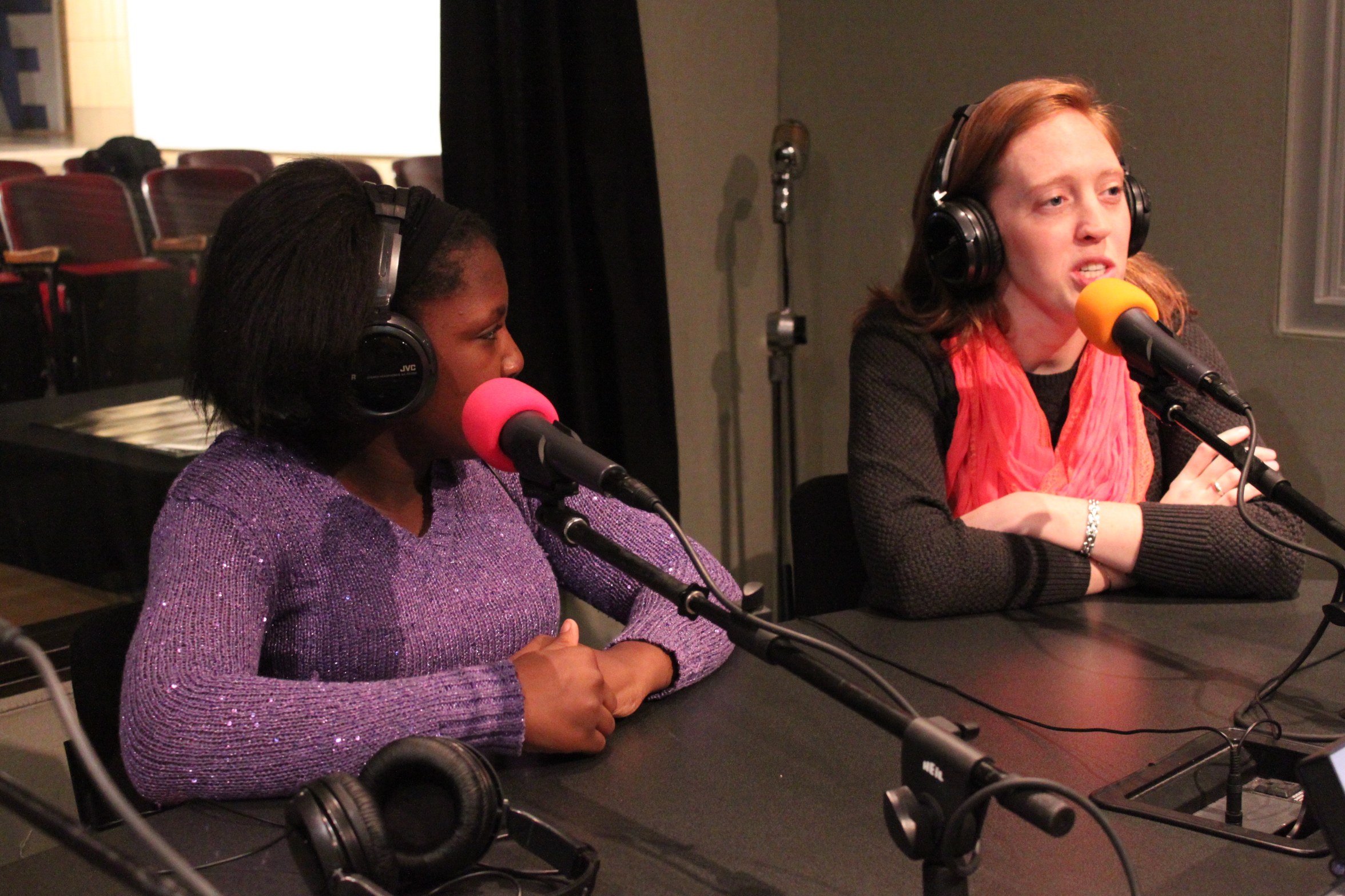 An adult and a youth wearing headphones speaking into microphones in a radio studio