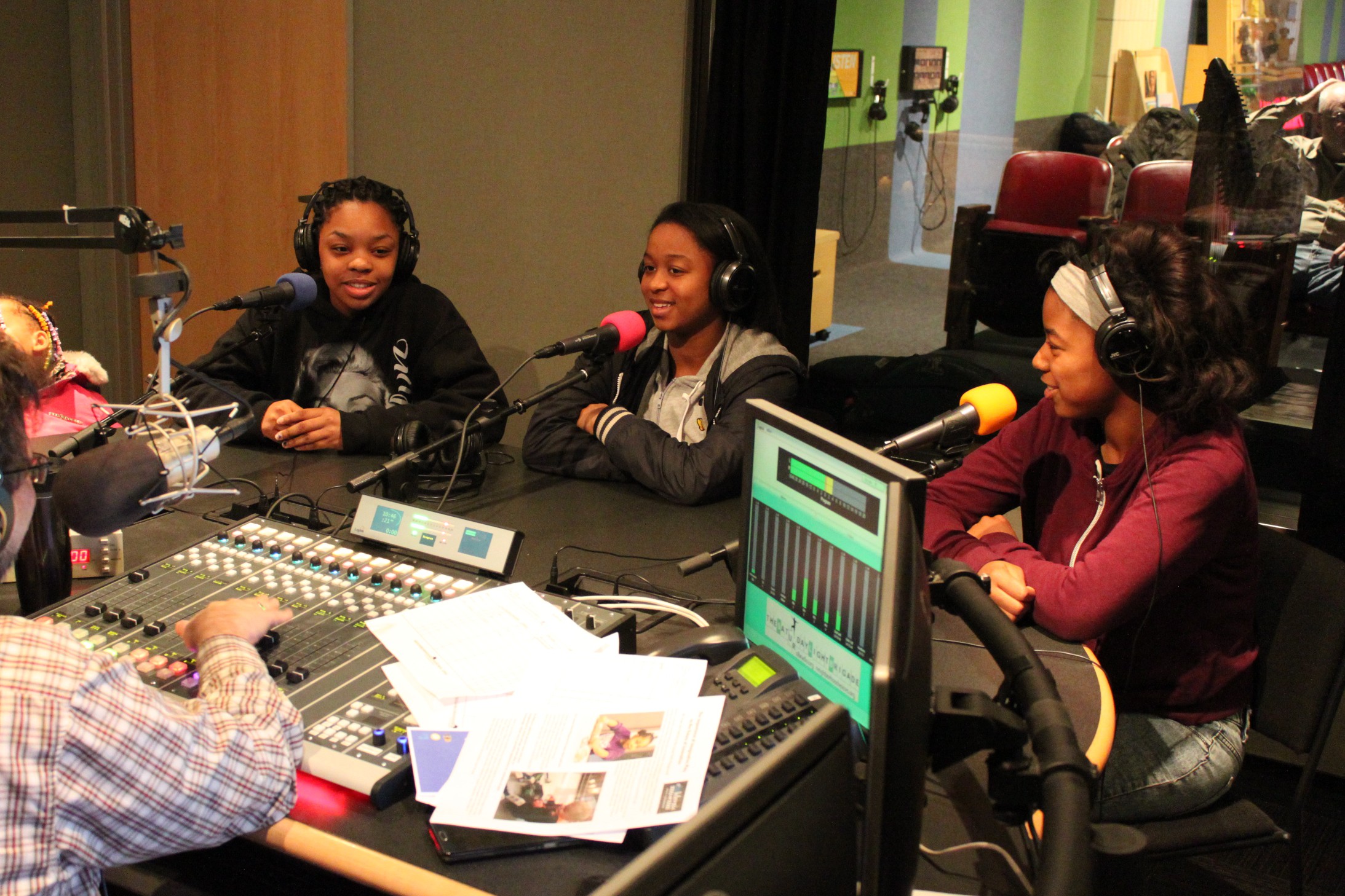 Three teens wearing headphones speaking into microphones being interviewed by an adult wearing headphones sitting behind a microphone and a mixing board in a radio studio