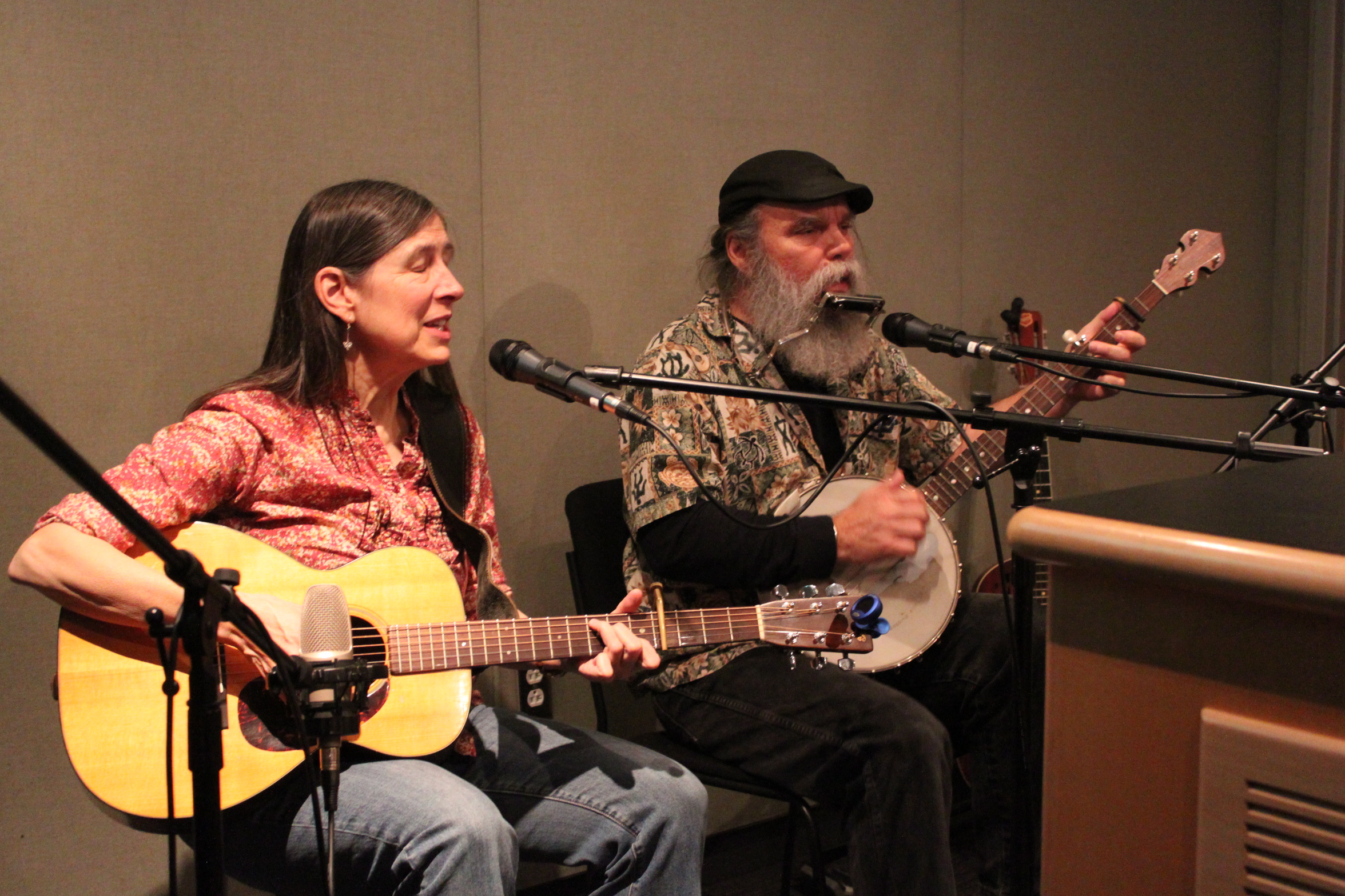 An adult playing acoustic guitar and an adult playing banjo, both singing into microphones in a radio studio