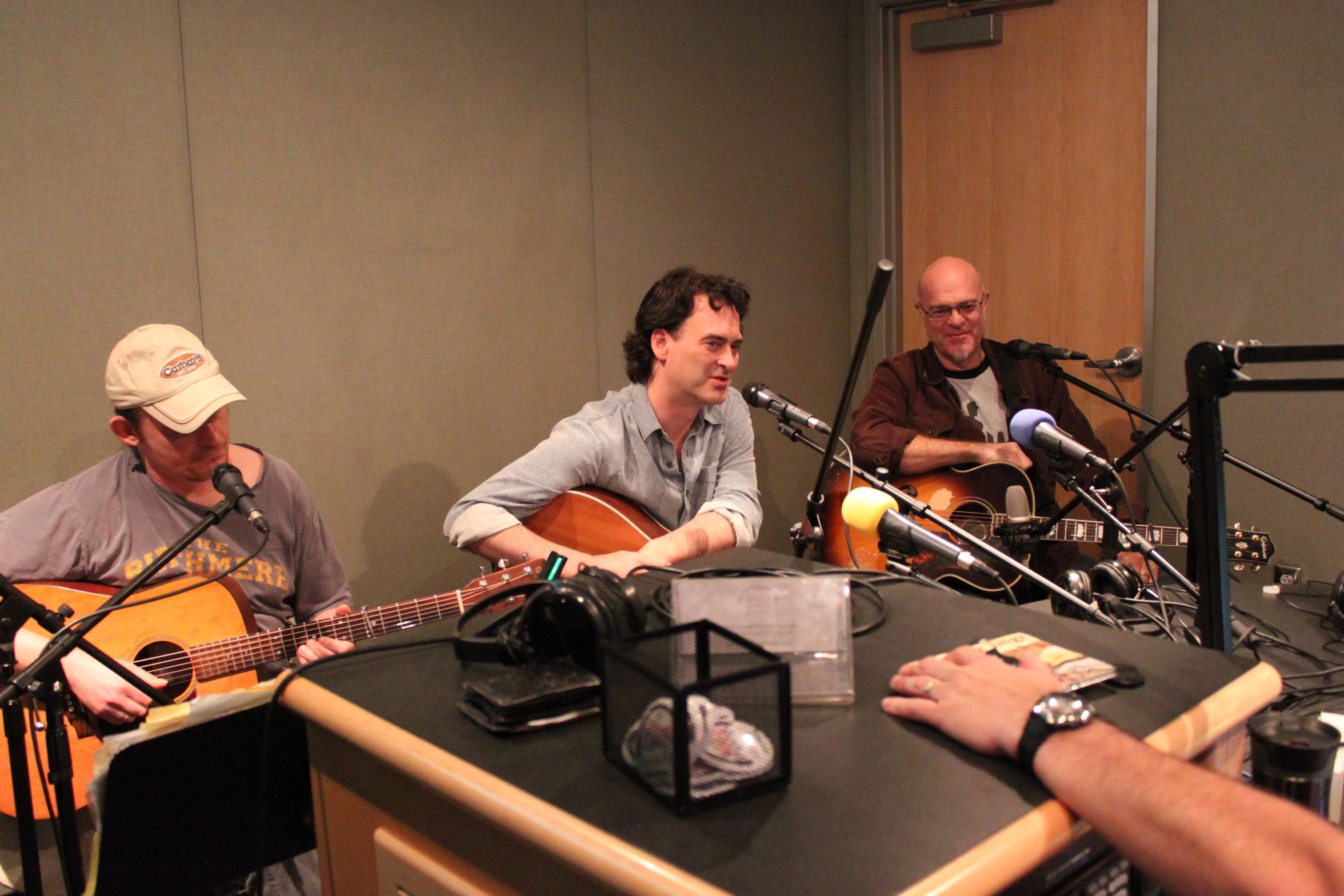 Three adults playing acoustic guitar and singing into microphones in a radio studio
