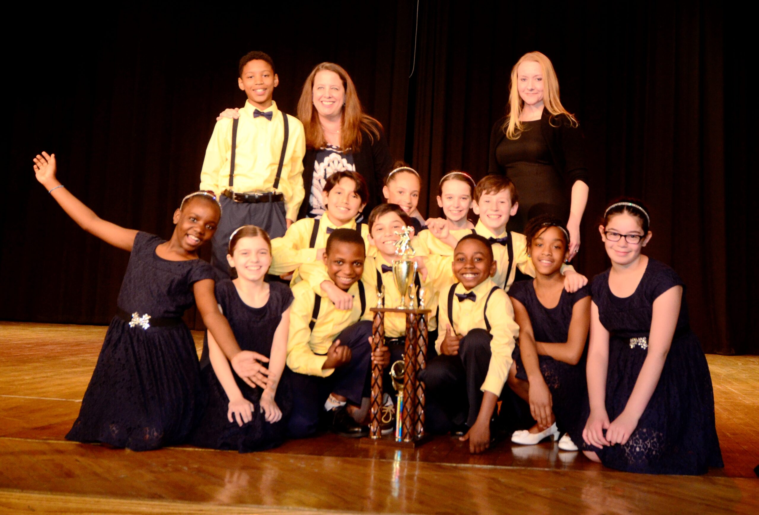 Eleven youth sitting and a youth and two adults standing behind a trophy and smiling on a stage
