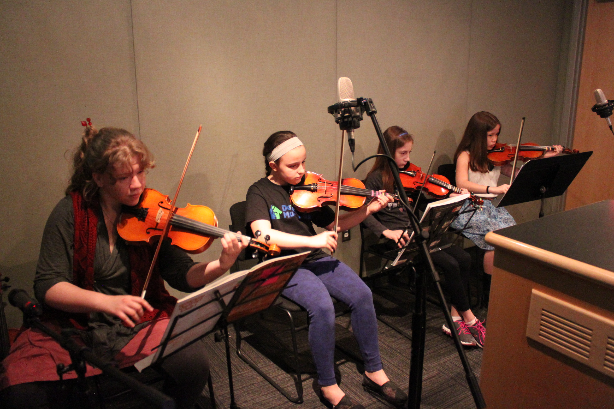 Three youth playing violins and one youth playing viola looking at sheet music on music stands in a radio studio