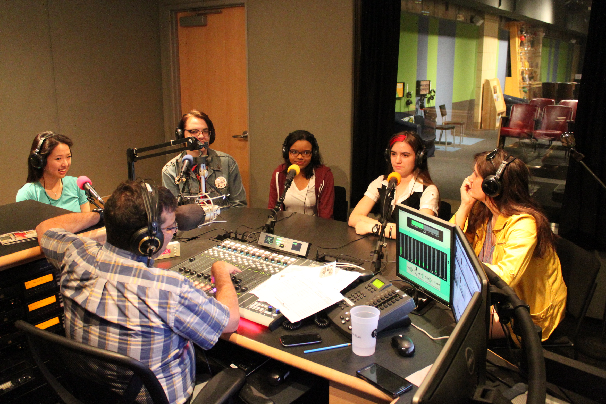 Five adults wearing headphones speaking into microphones being interviewed by an adult wearing headphones sitting behind a microphone and a mixing board in a radio studio