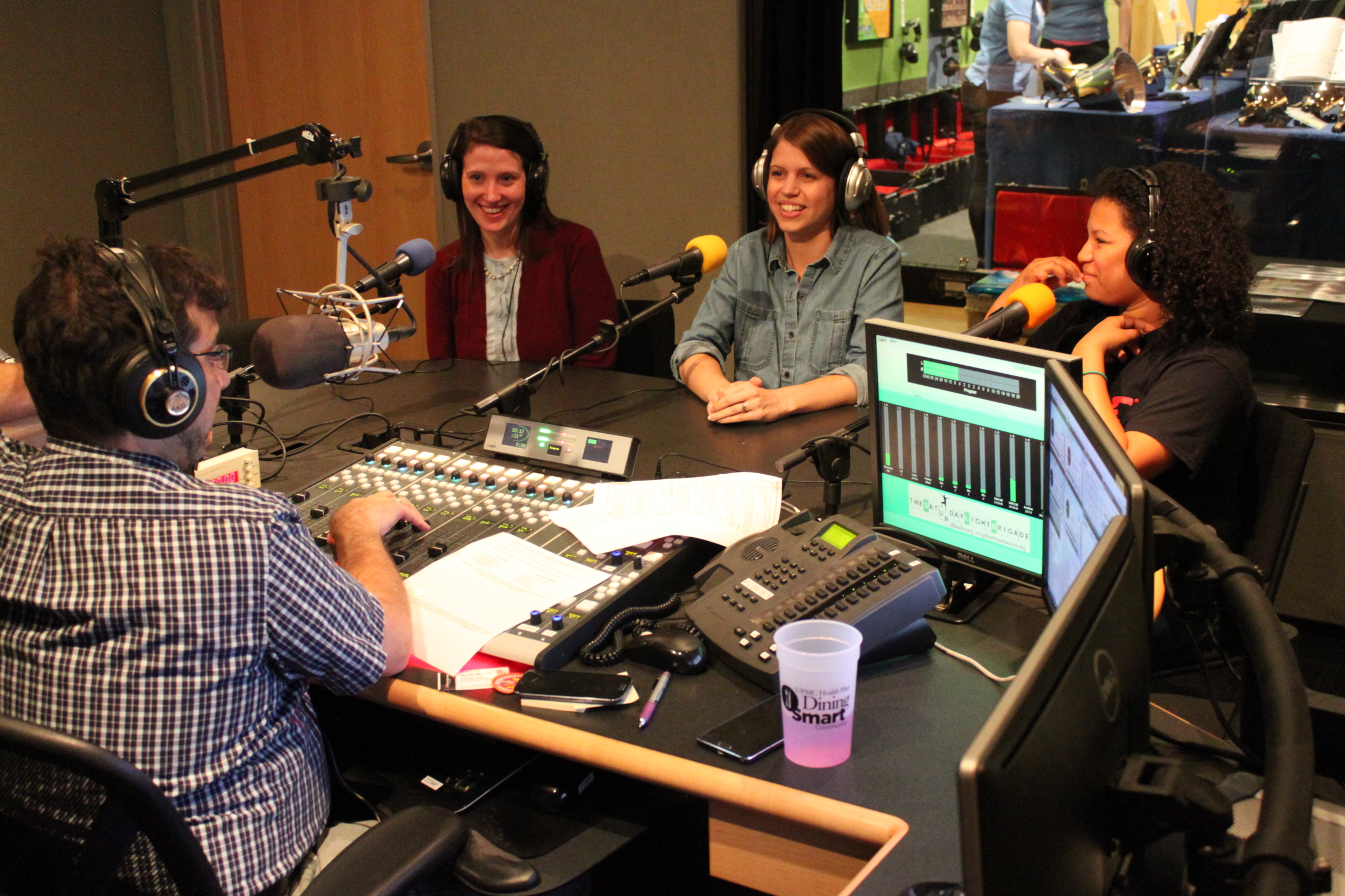 A youth and two adults wearing headphones speaking into microphones being interviewed by an adult wearing headphones sitting behind a microphone and a mixing board in a radio studio