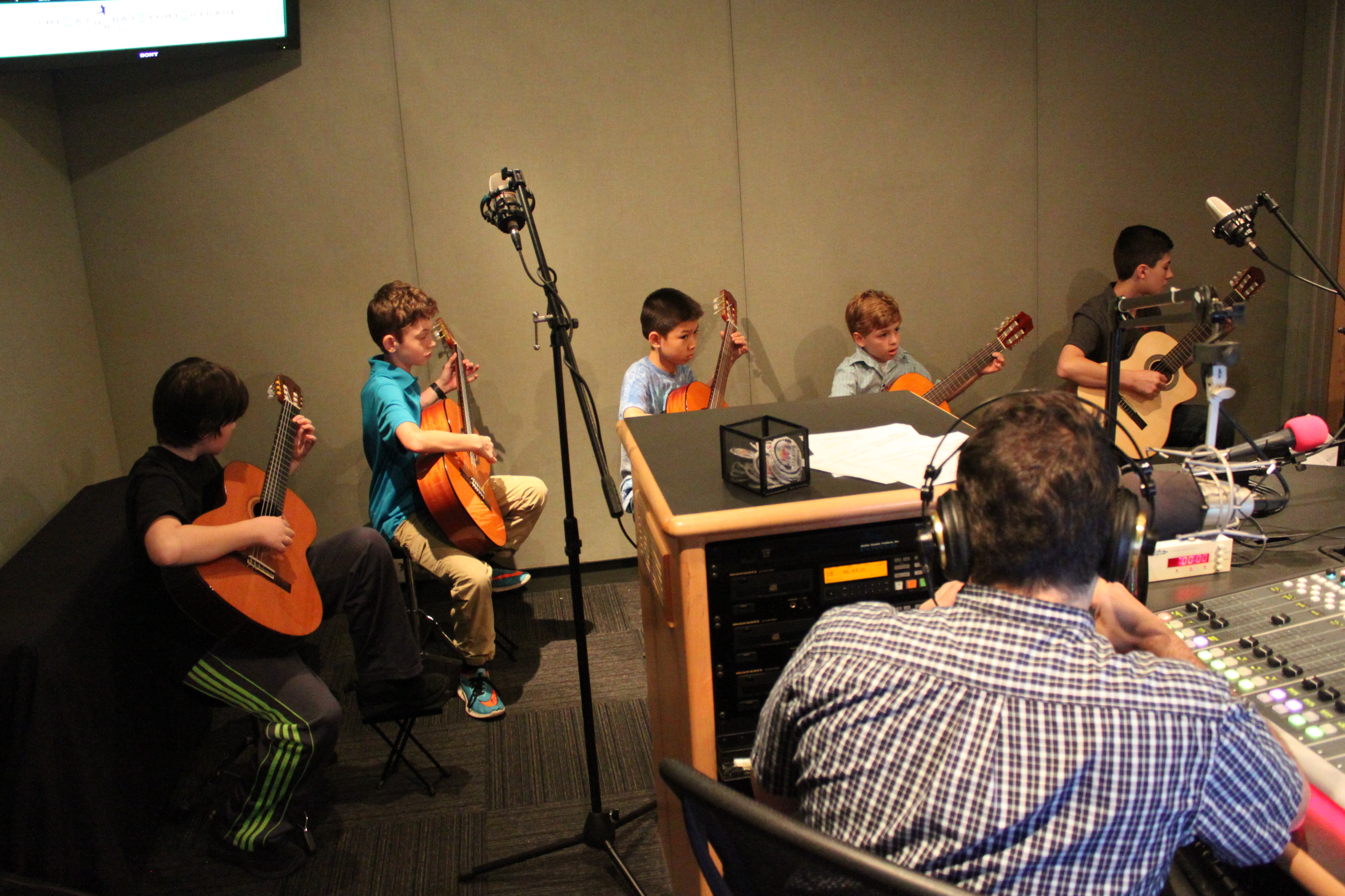 Five youth playing acoustic guitars and an adult wearing headphones sitting behind a mixing board listening in a radio studio