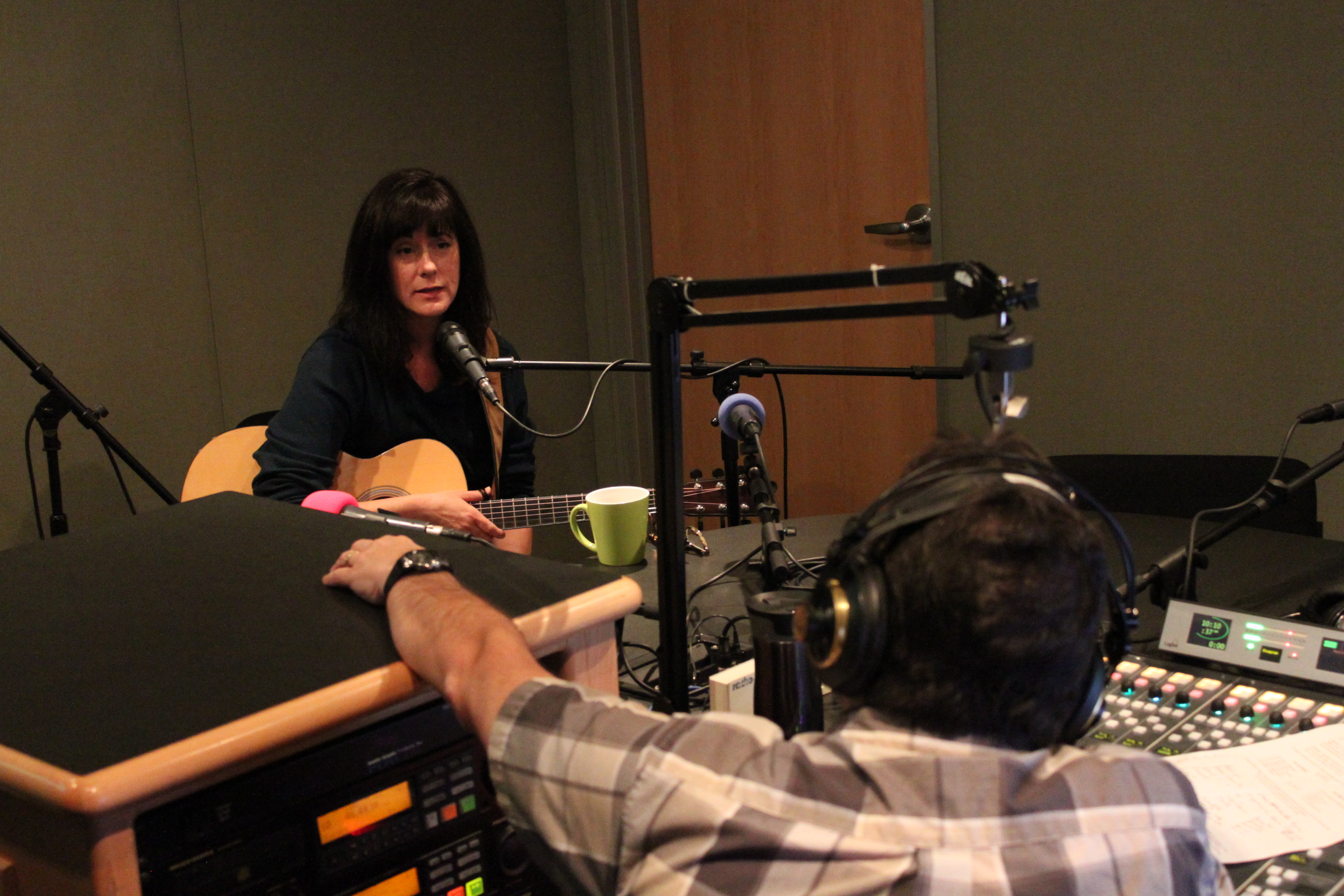 An adult holding an acoustic guitar sitting behind a microphone being interviewed by an adult wearing headphones sitting behind a microphone and a mixing board in a radio studio