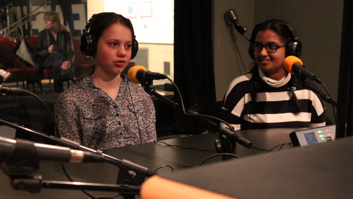 Two teens wearing headphones speaking into microphones in a radio studio