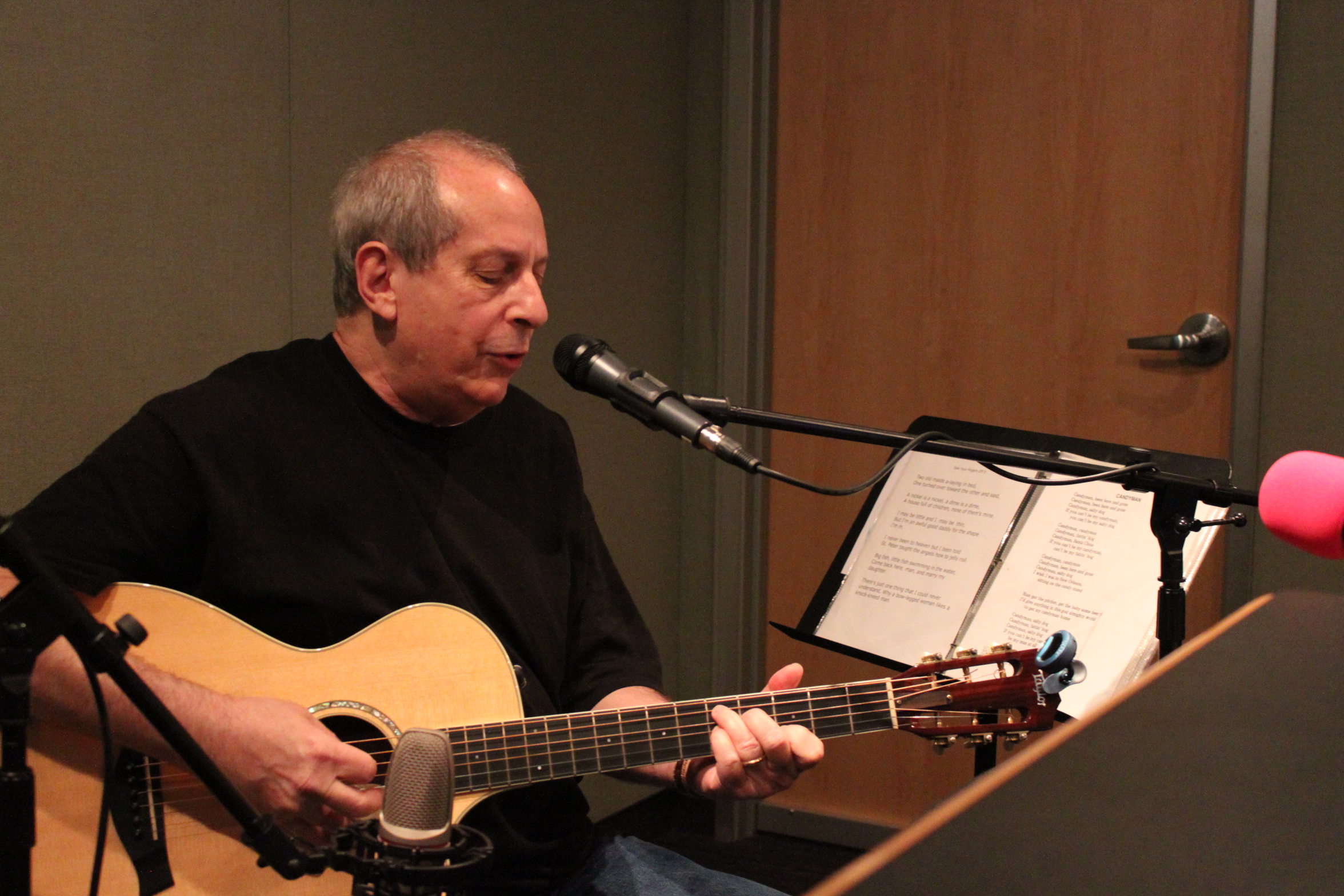 An adult playing an acoustic guitar singing into a microphone in a radio studio