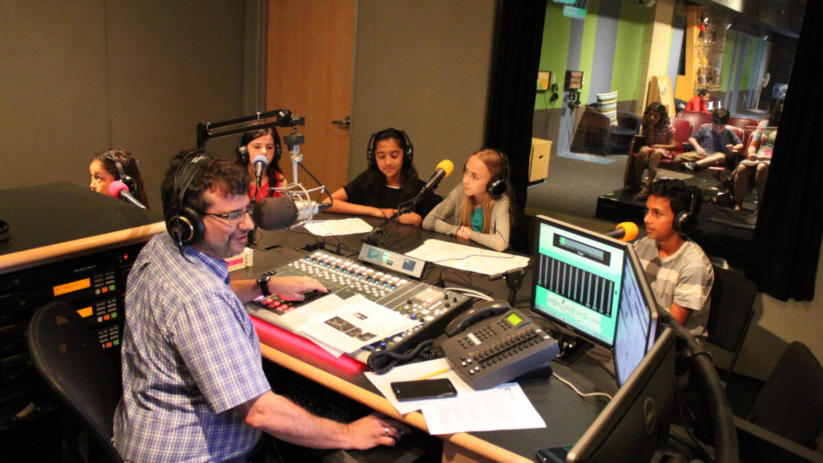 Four youth wearing headphones sitting behind microphones being interviewed by an adult wearing headphones sitting behind a microphone and a mixing board in a radio studio
