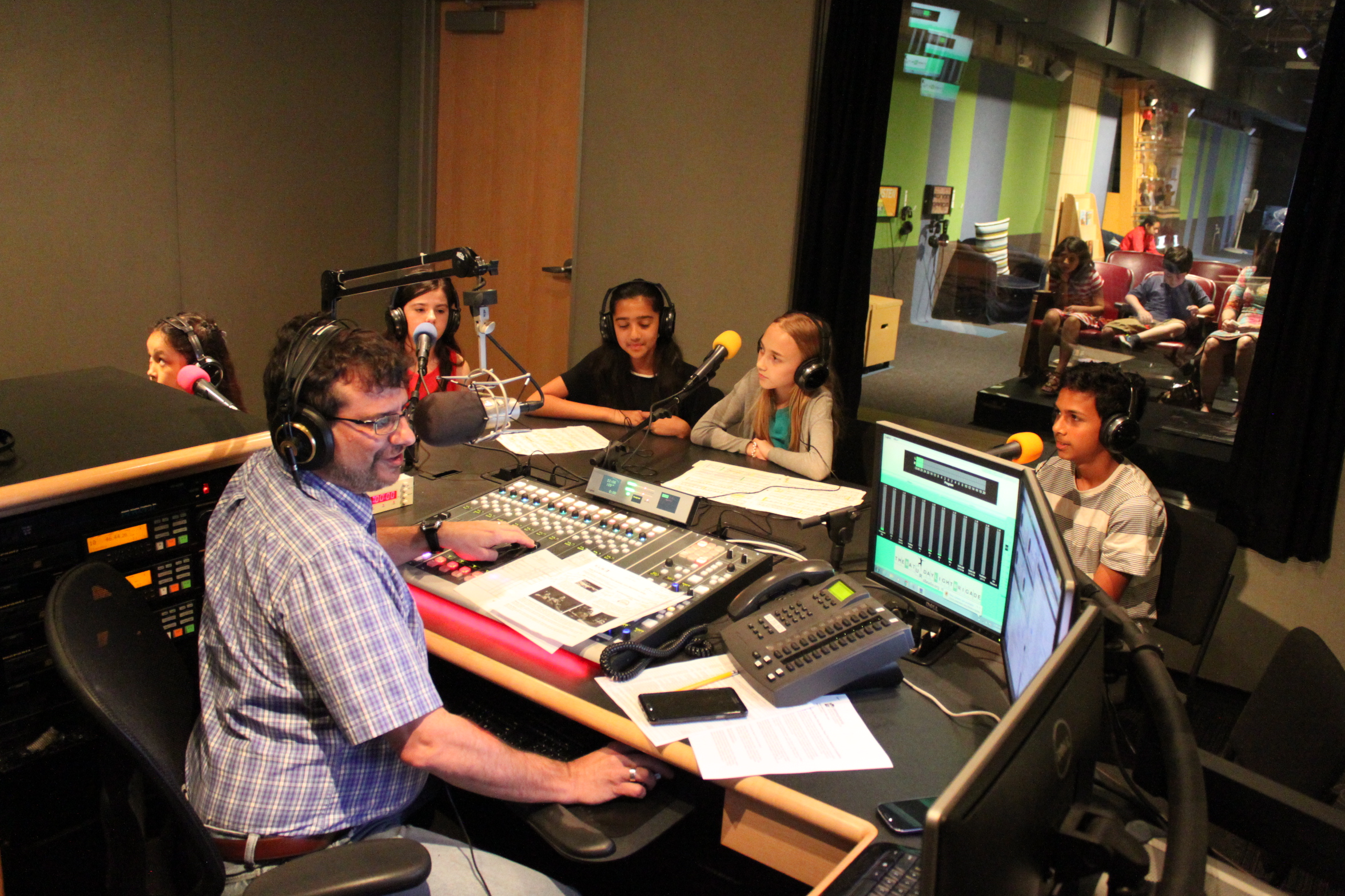Four youth wearing headphones sitting behind microphones being interviewed by an adult wearing headphones sitting behind a microphone and a mixing board in a radio studio