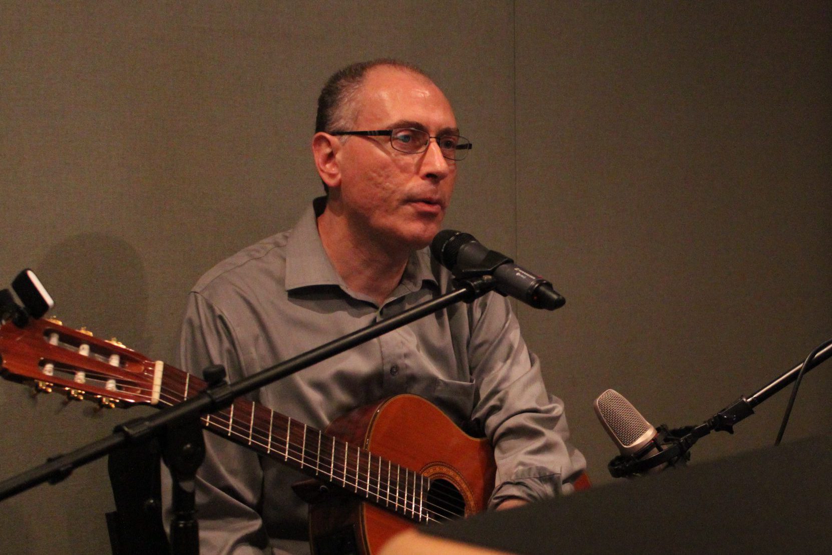 An adult holding an acoustic guitar sitting behind microphones in a radio studio