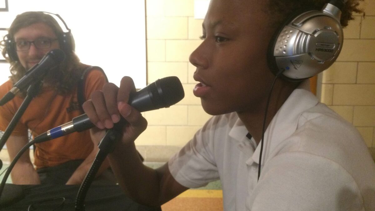 An adult wearing headphones sitting behind a microphone and a youth wearing headphones speaking into a microphone